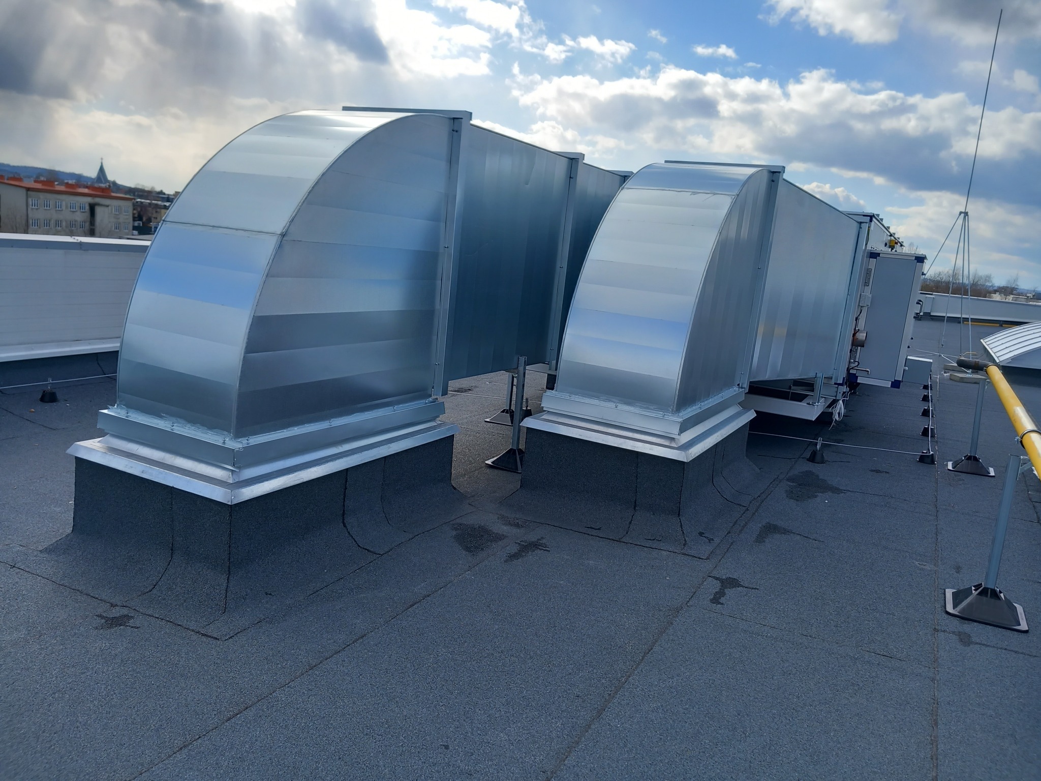 System kanałów wentylacyjnych z ocynkowanej blachy na dachu budynku, zabezpieczony odgromnik, widok na miasto w tle pod błękitnym niebem