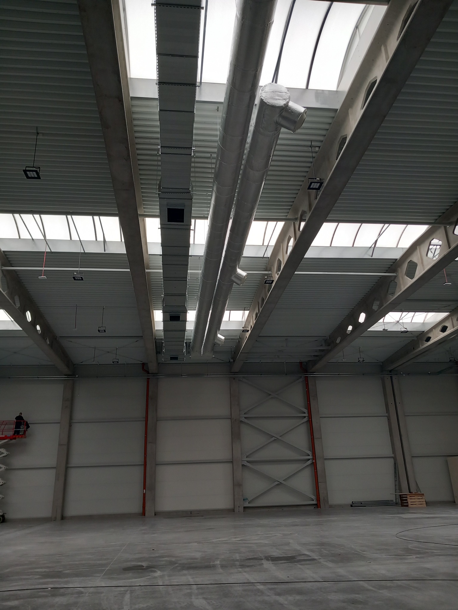 Wnętrze hali przemysłowej z widoczną instalacją wentylacyjną pod sufitem, pracownik na podnośniku koszowym w lewym dolnym rogu.