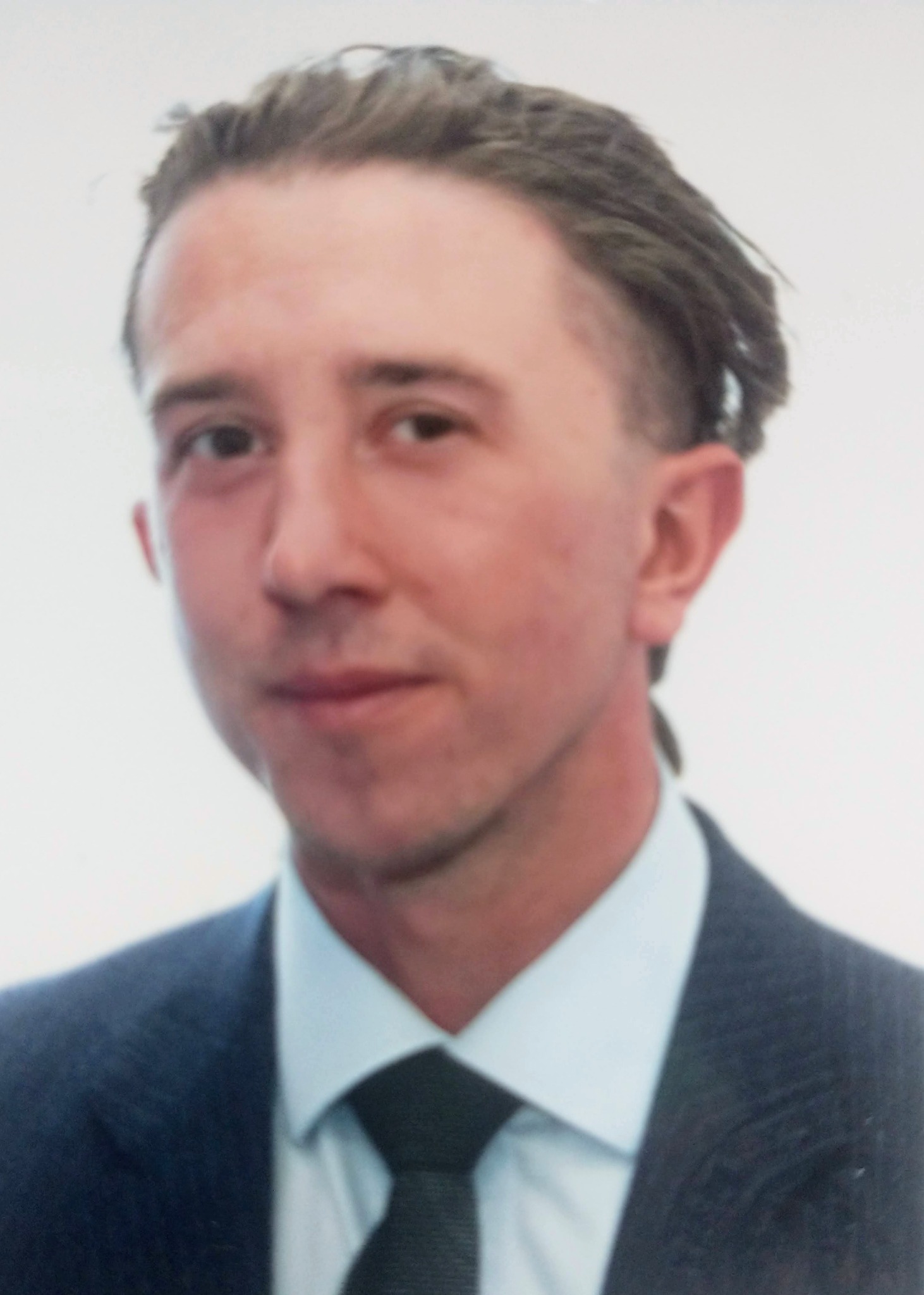 Portret mężczyzny w ciemnym garniturze i krawacie, z zaczesanymi do tyłu włosami, na jasnym tle.