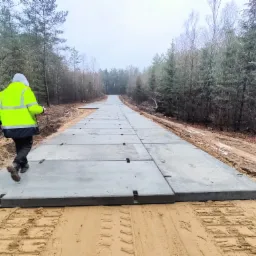 Robocza droga tymczasowa ułożona z betonowych płyt na piaszczystym terenie budowy, z pracownikiem w odblaskowej kurtce idącym wzdłuż niej w kierunku lasu w tle.