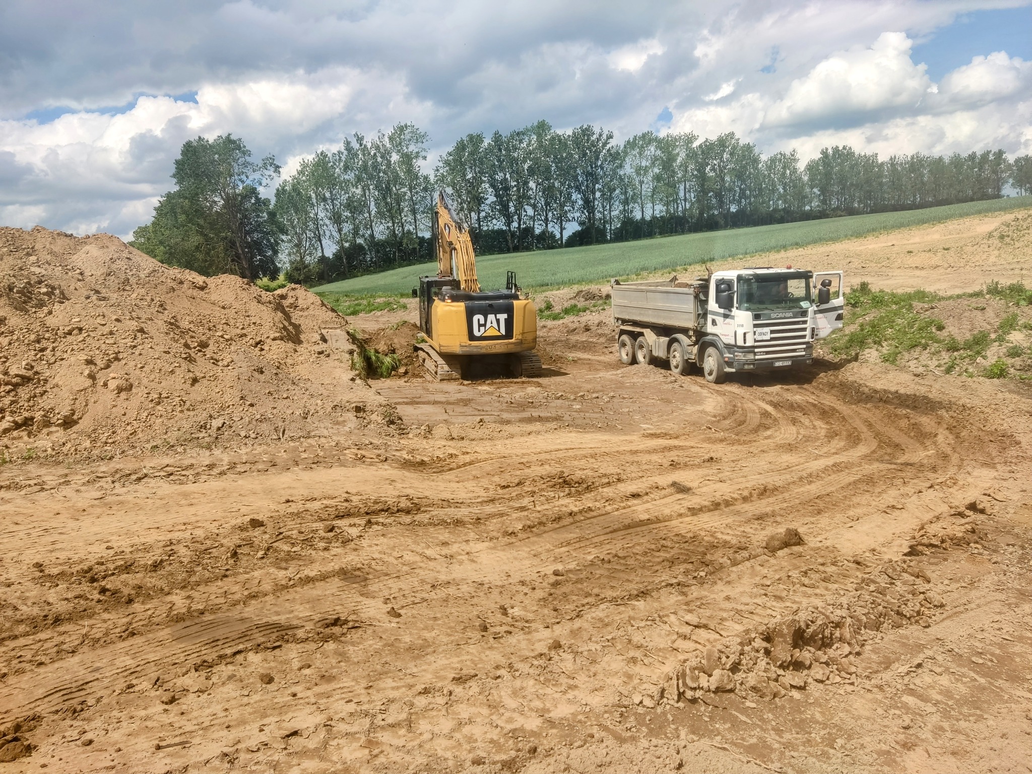 Żółta koparka CAT i biała wywrotka Scania pracują na placu budowy z dużą hałdą ziemi, w tle linia drzew i pole pod pochmurnym niebem.