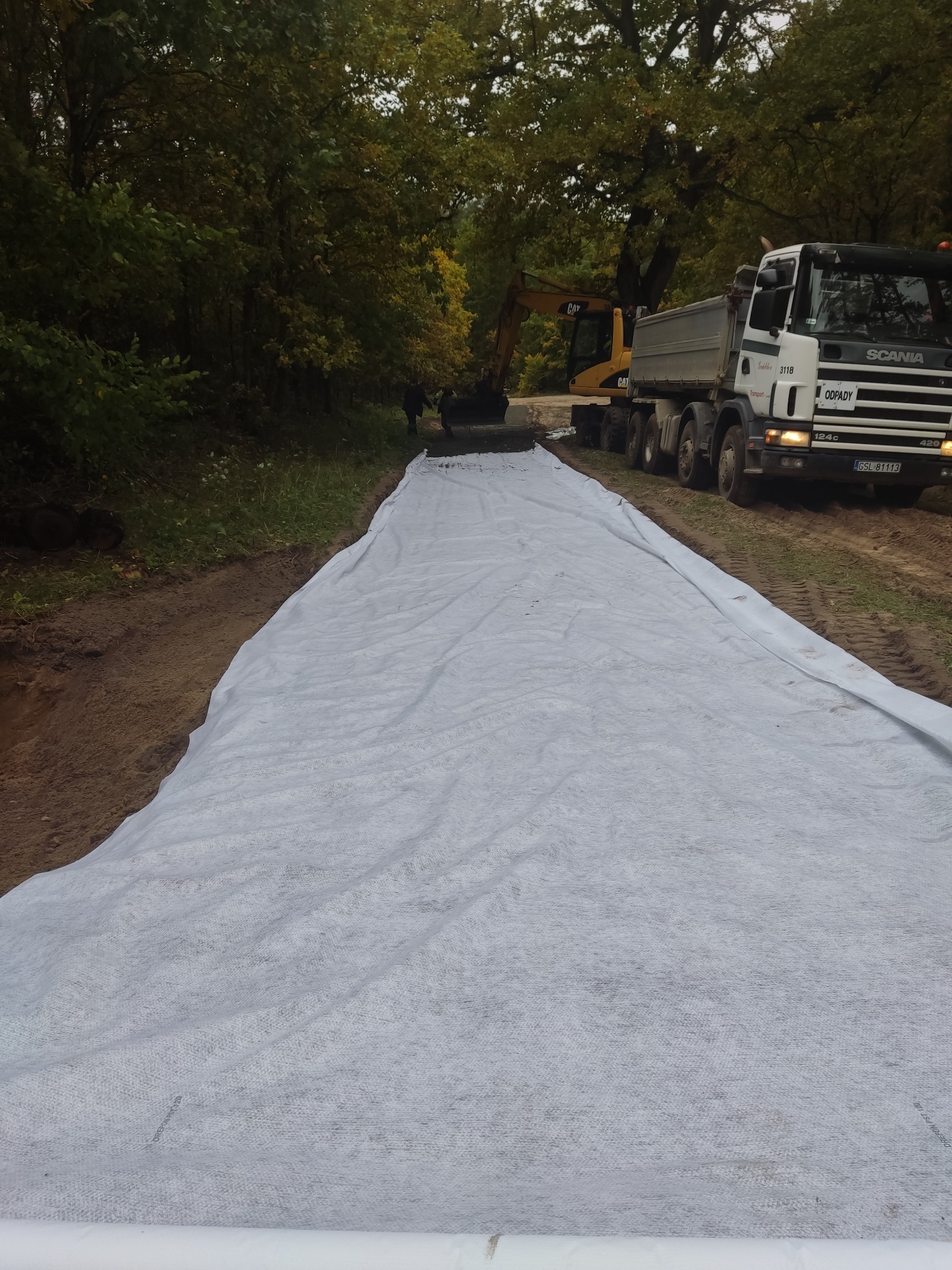 Rozkładanie geowłókniny na gruncie pod budowę drogi, w tle koparka i wywrotka Scania