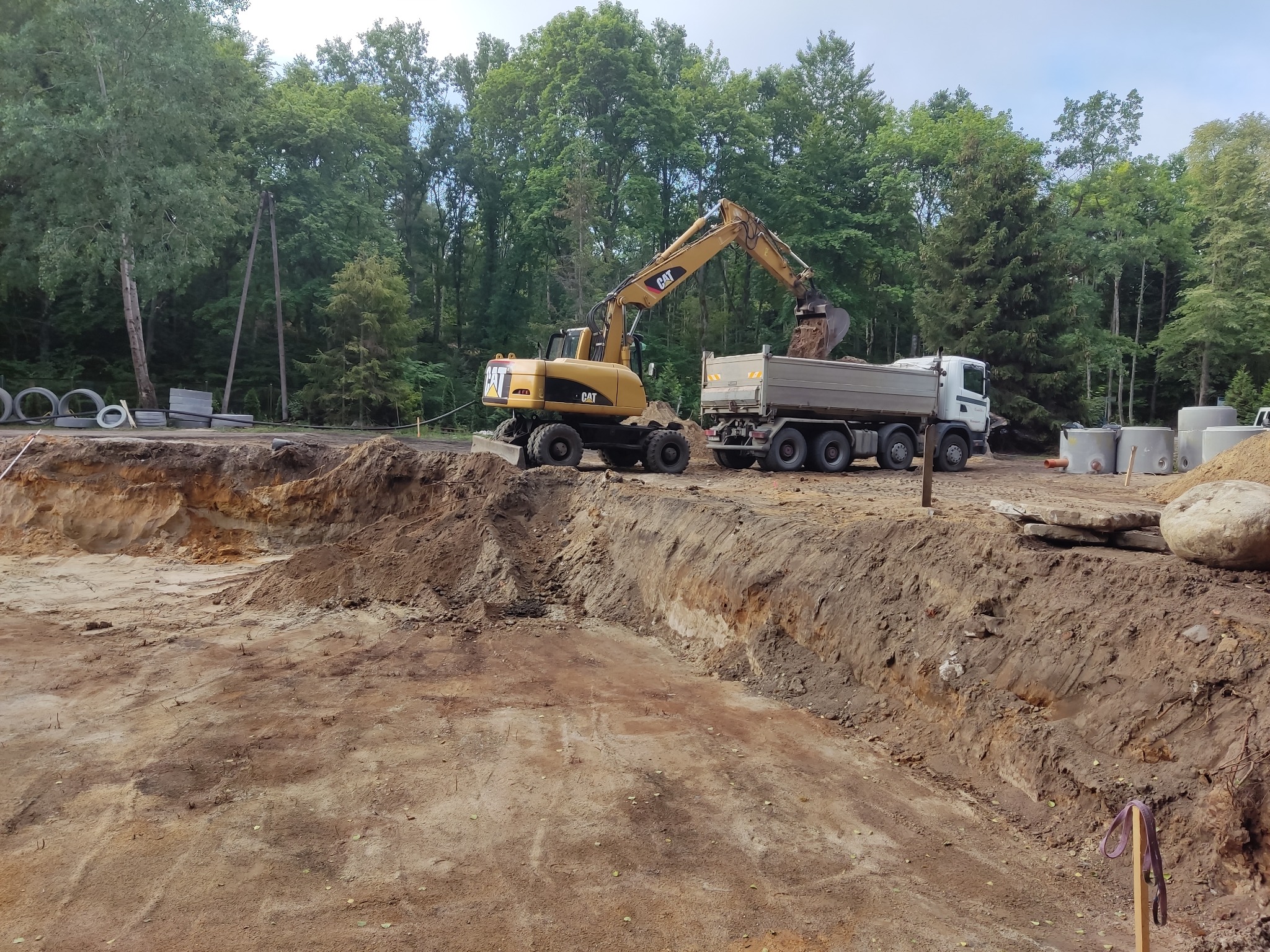 Żółta koparka CAT ładuje ziemię do białej ciężarówki na tle wykopu, stosu betonowych kręgów i zieleni drzew.