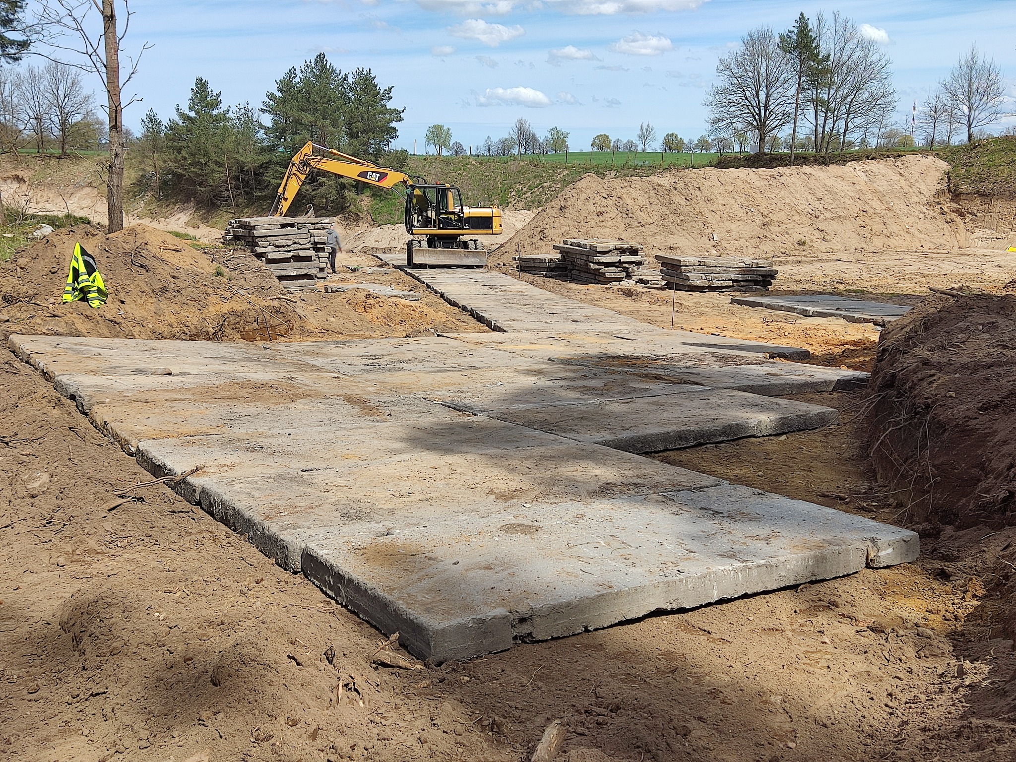 Żółta koparka CAT na placu budowy z ułożonymi betonowymi płytami, w tle hałda ziemi i drzewa.