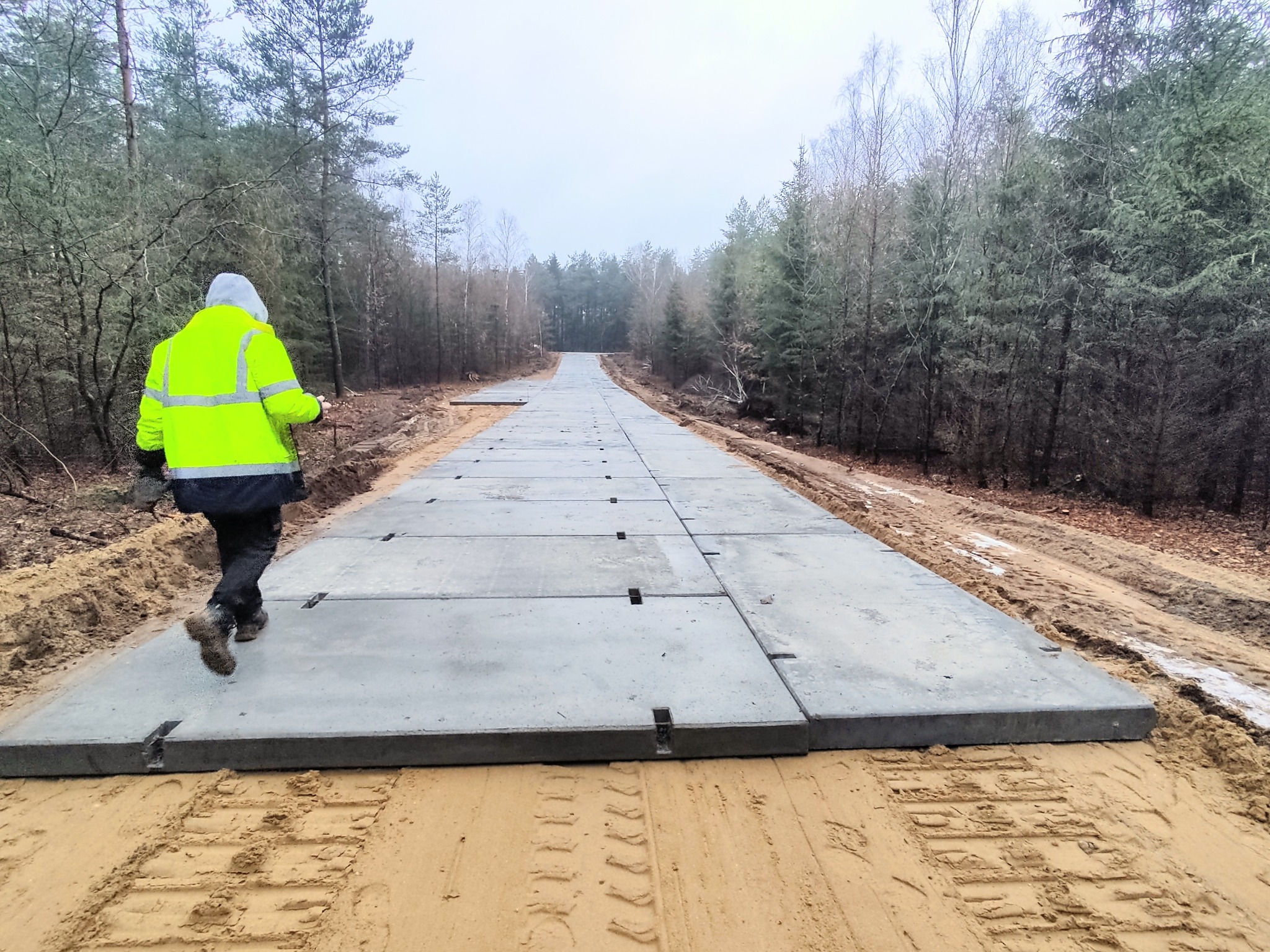 Robocza droga tymczasowa ułożona z betonowych płyt na piaszczystym terenie budowy, z pracownikiem w odblaskowej kurtce idącym wzdłuż niej w kierunku lasu w tle.