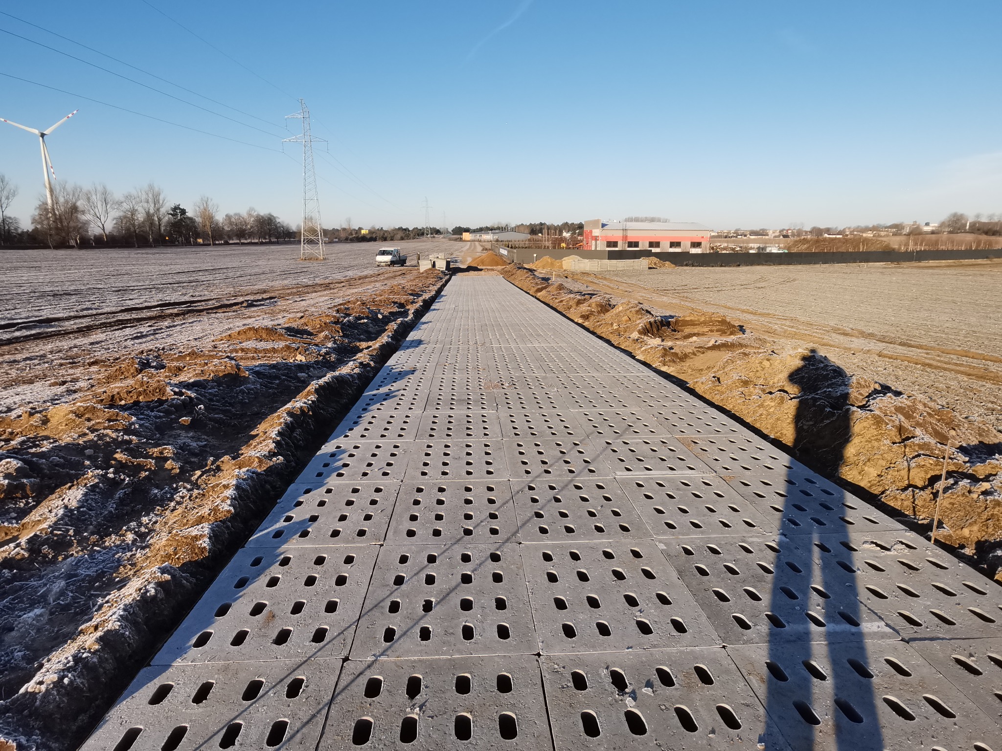 Szara, ażurowa nawierzchnia tymczasowa ułożona na wykopie w polu, w tle widoczny budynek przemysłowy, turbina wiatrowa i słupy wysokiego napięcia pod błękitnym niebem.