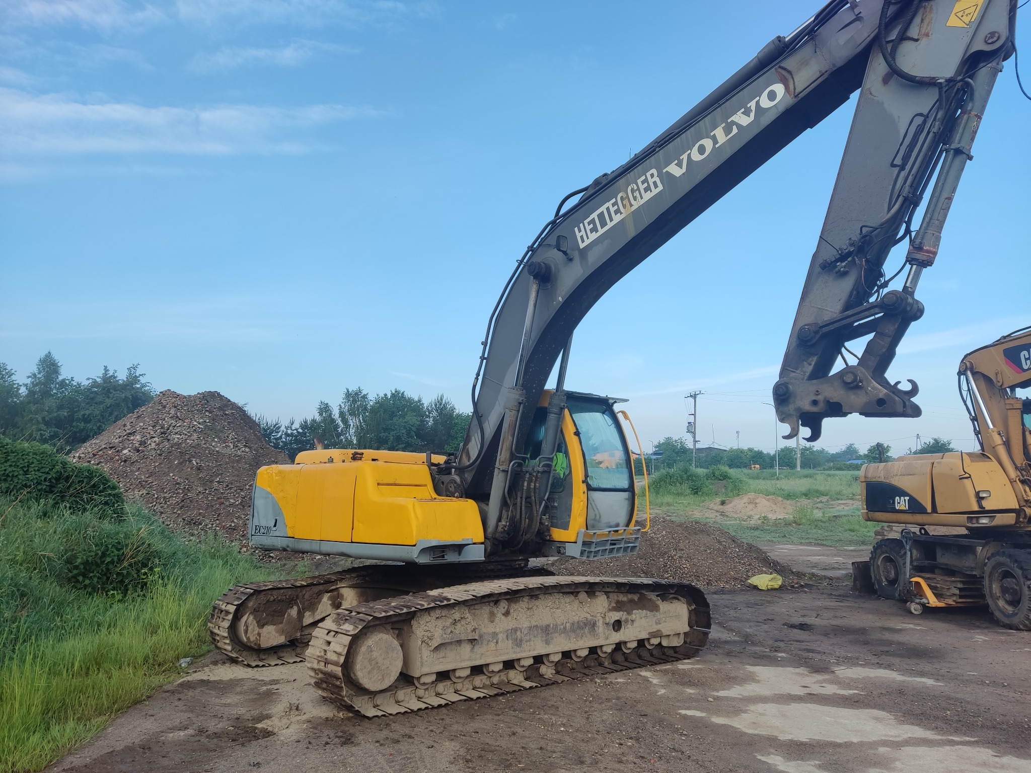 Żółto-szara koparka Volvo Hettegger na placu budowy, obok sterta gruzu i druga maszyna CAT, pochmurne niebo w tle.