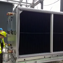 Dwóch monterów w kaskach i kamizelkach odblaskowych instaluje duży, modułowy system wentylacyjny na zewnątrz budynku, widoczne przyłącza rurowe.