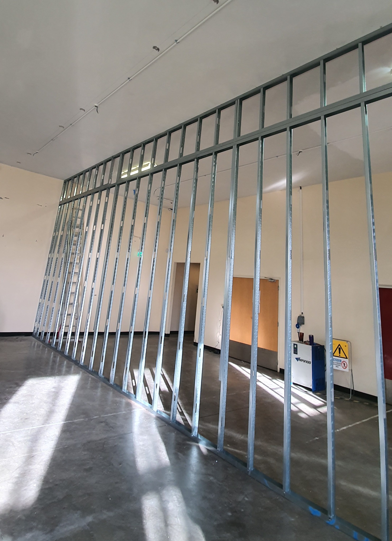 Stelaż metalowy ściany działowej w trakcie montażu w przestronnym pomieszczeniu, widoczne drzwi w tle i sprzęt budowlany.