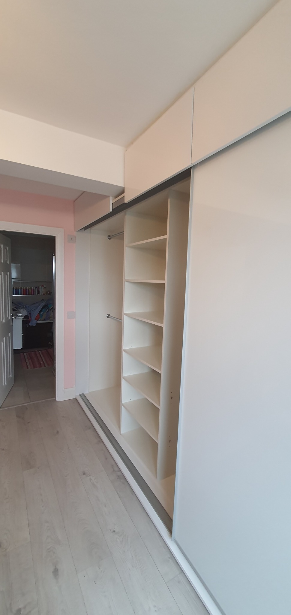 Biała szafa wnękowa z przesuwnymi drzwiami, widoczne półki wewnątrz, jasne drewniane panele na podłodze, w tle otwarte drzwi do innego pokoju z różowymi ścianami.