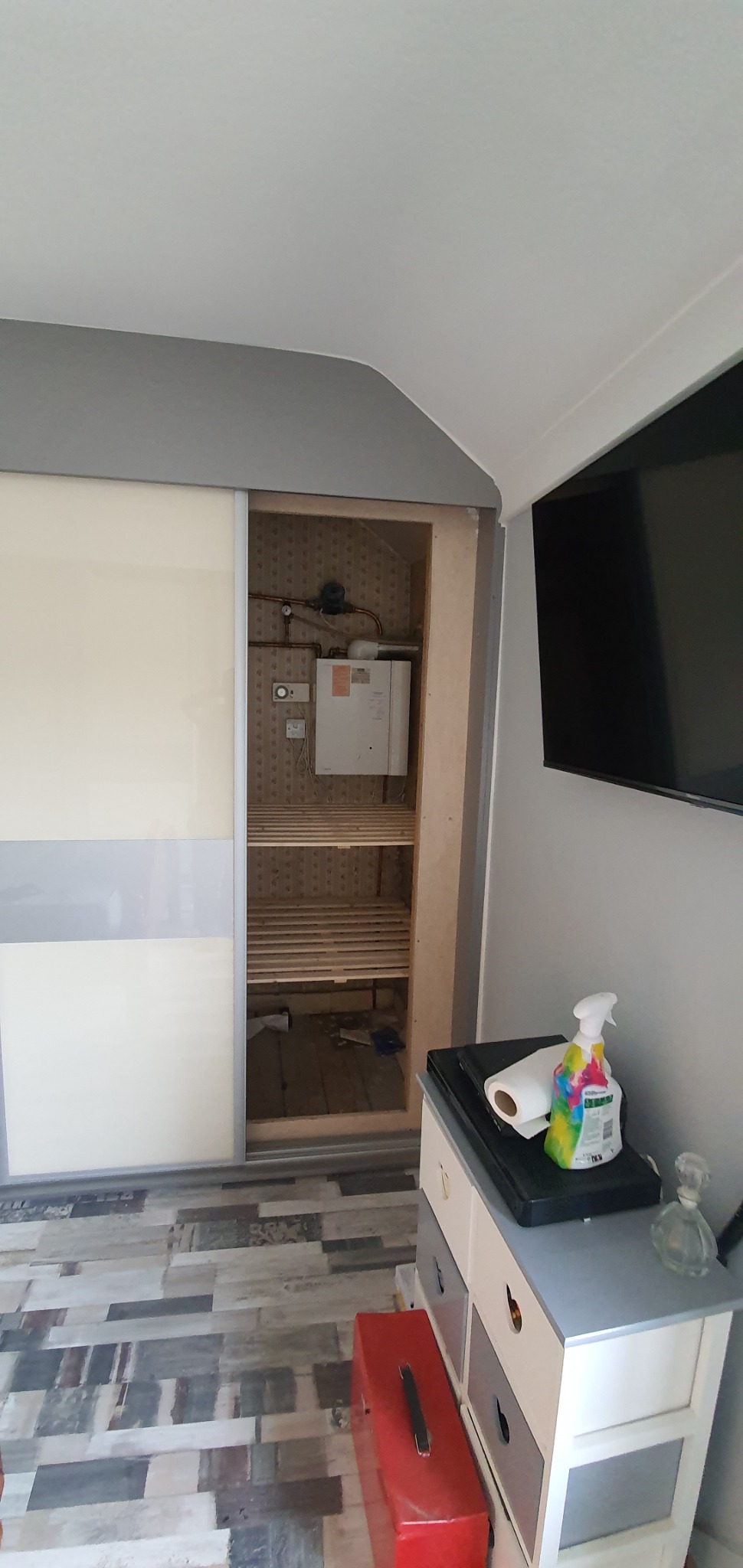 Wnętrze szafy wnękowej z przesuwnymi drzwiami w trakcie montażu, widoczne półki i instalacja grzewcza na ścianie, obok komoda z akcesoriami do czyszczenia.