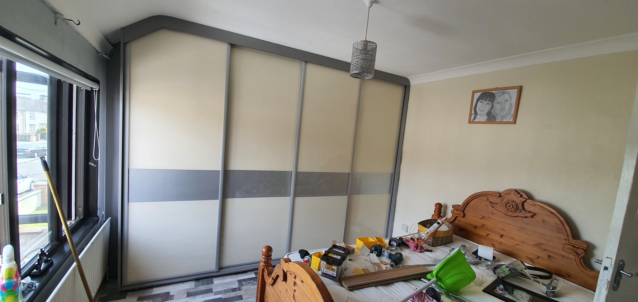 Duża szafa przesuwna z beżowymi i szarymi panelami w sypialni, widoczny zagracony łóżko i okno.