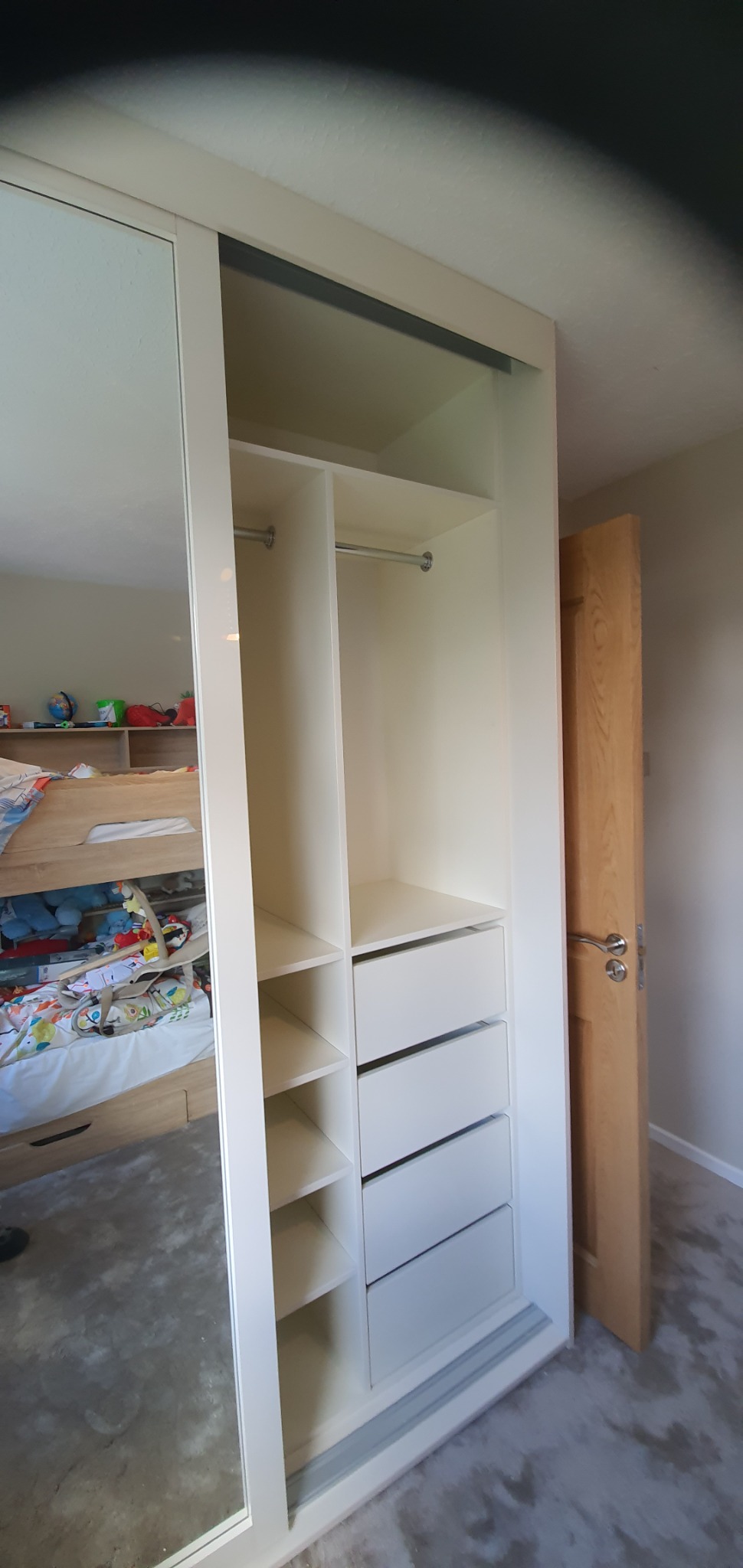 Wnętrze białej szafy wnękowej z lustrzanymi drzwiami przesuwnymi, widoczne półki, szuflady i drążek na ubrania. W lustrze odbija się pokój dziecięcy z łóżkiem piętrowym i zabawkami.
