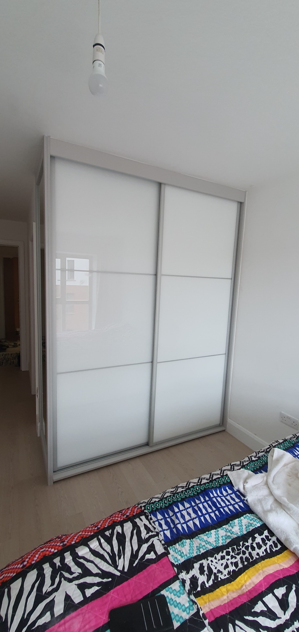 Biała szafa przesuwna z czterema panelami na frontach, z widocznym odbiciem okna, stojąca w sypialni z kolorową narzutą na łóżku i wiszącą żarówką.