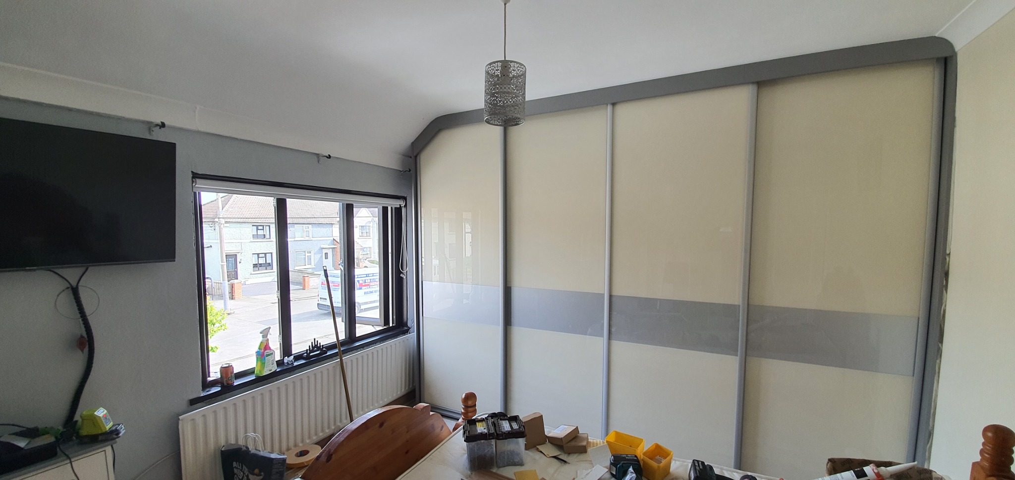 Jasna szafa przesuwna z szarymi elementami dekoracyjnymi, widoczna w pokoju z oknem i wiszącą lampą, podczas montażu.