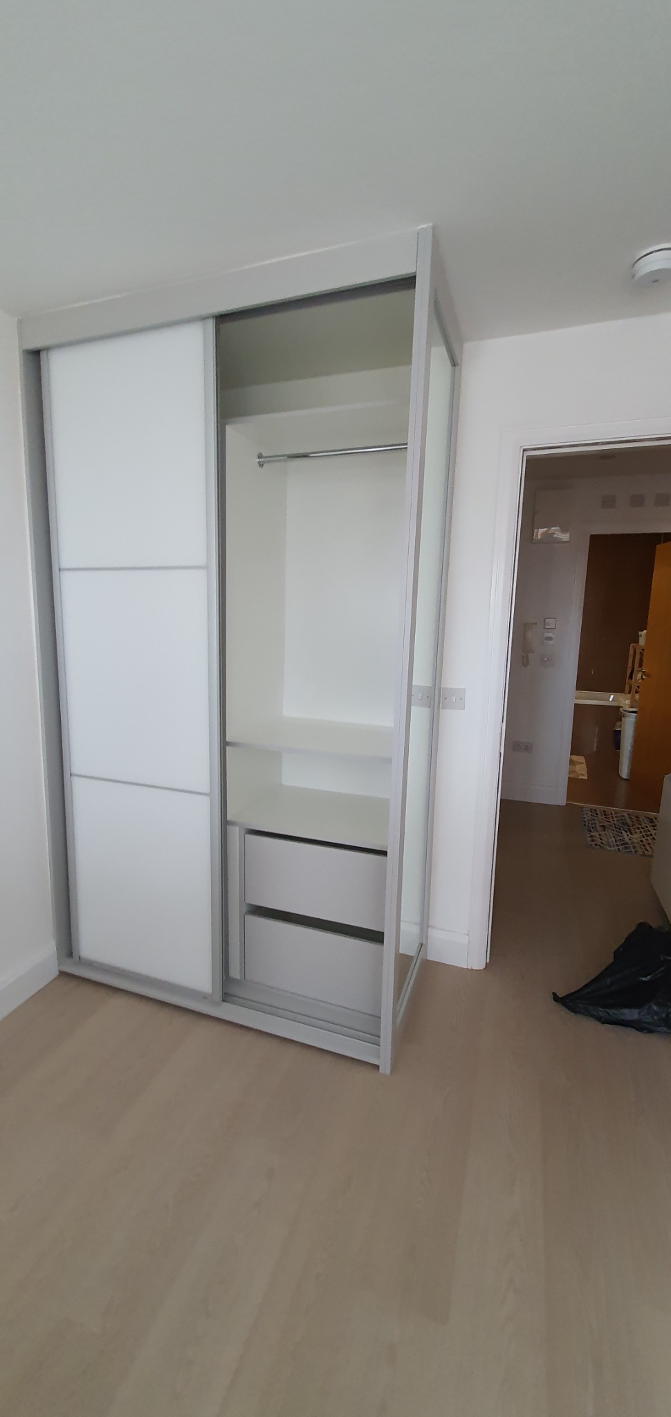 Wnętrze mieszkania z białą szafą wnękową z przesuwnymi drzwiami, częściowo otwartą, ukazującą półki i szuflady; widoczny fragment łazienki w tle.