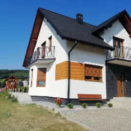 Dwupiętrowy dom z białą elewacją, fragmentami w kolorze drewna i czarną dachówką na tle błękitnego nieba; widoczny fragment ogrodu z basenem i placem zabaw.