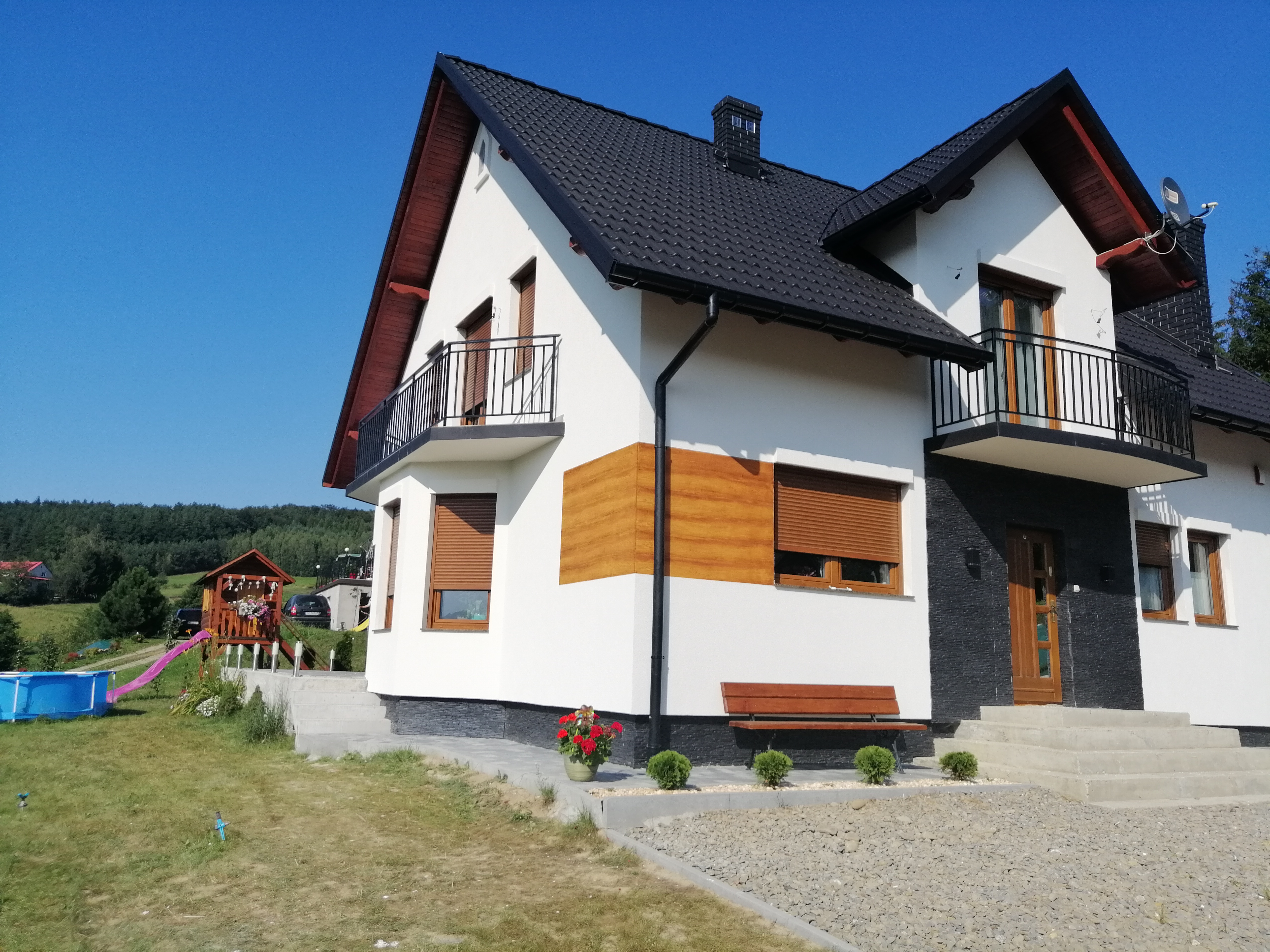 Dwupiętrowy dom z białą elewacją, fragmentami w kolorze drewna i czarną dachówką na tle błękitnego nieba; widoczny fragment ogrodu z basenem i placem zabaw.