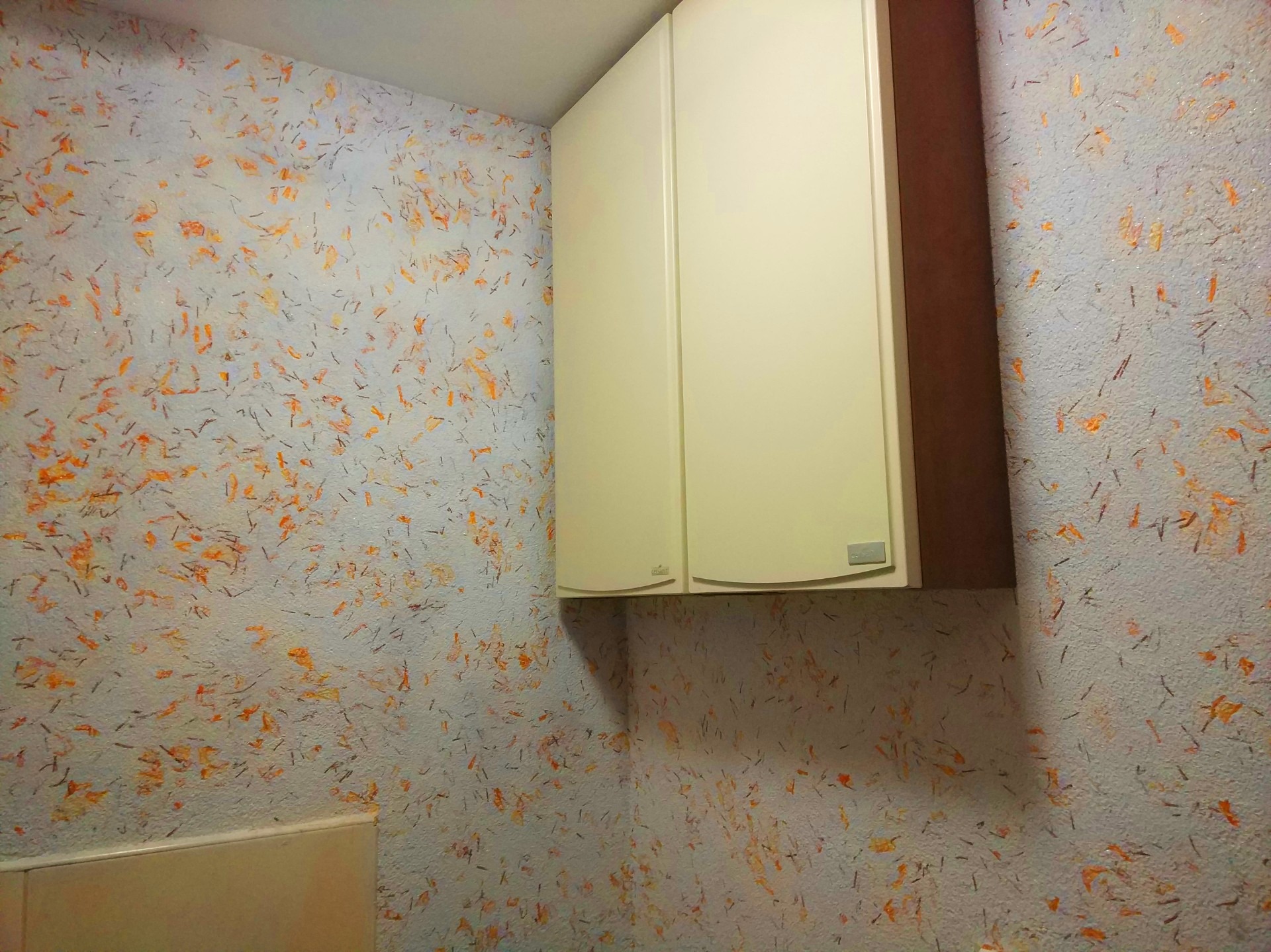 Ściana pokryta dekoracyjnym tynkiem strukturalnym z pomarańczowymi i brązowymi elementami, obok wisząca szafka w kolorze kremowym z brązowym bokiem.