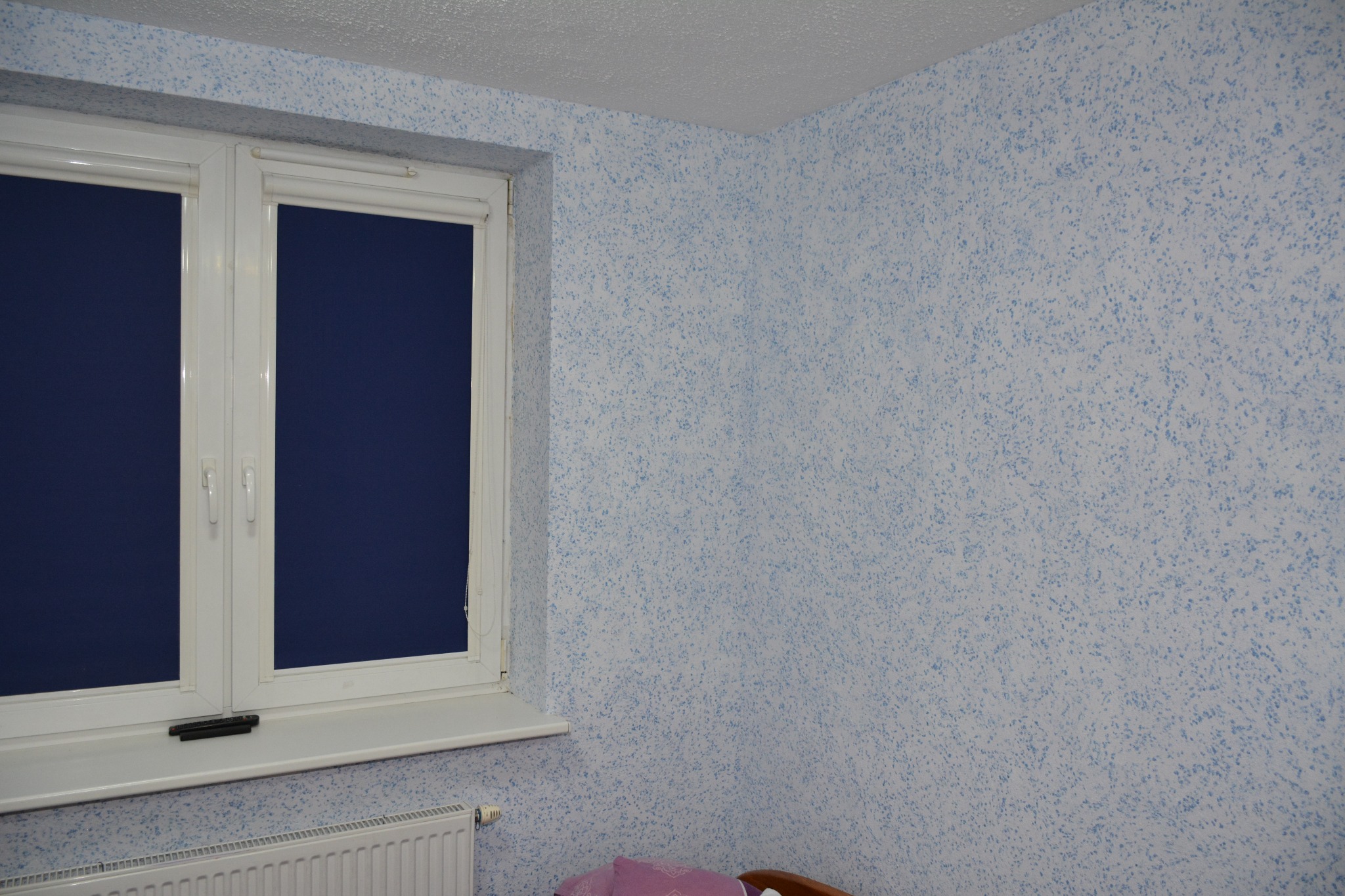 Pokój po remoncie: ściany pokryte jasnym tynkiem strukturalnym z niebieskimi akcentami, białe okno z zasłoniętymi roletami w kolorze granatowym, widoczny fragment grzejnika i łóżka.