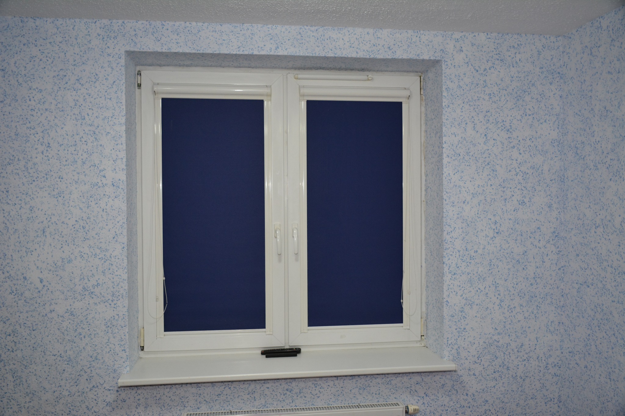 Pokój z oknem PCV i zasłoniętymi roletami w kolorze granatowym, ściany pokryte tynkiem strukturalnym w odcieniach bieli i błękitu.