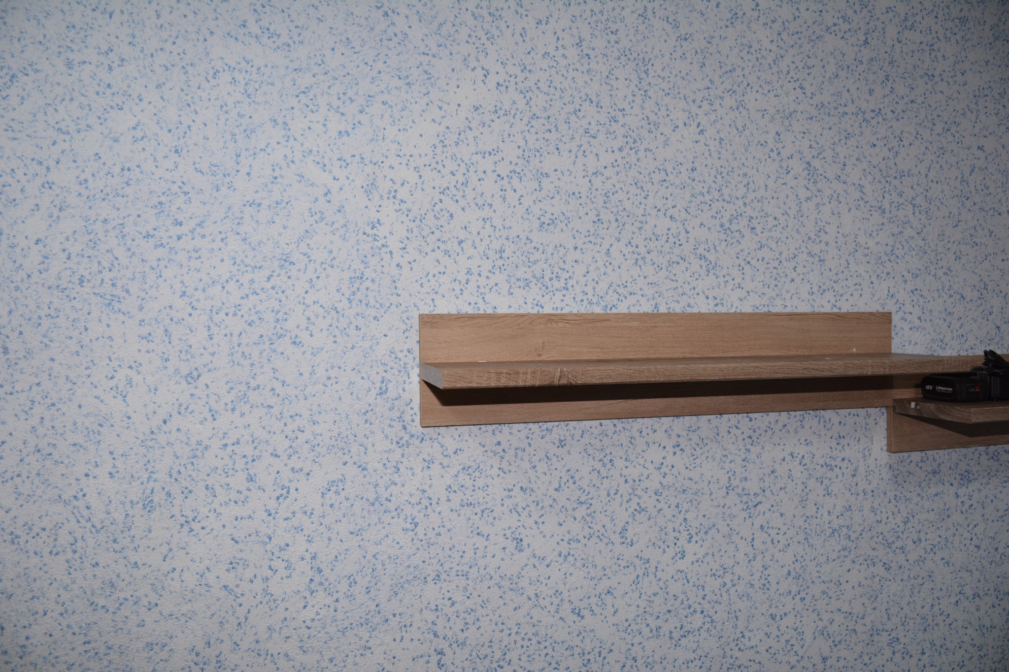 Jasnoniebieska ściana z fakturą kropek, drewniana półka z zamontowanym akumulatorem do elektronarzędzi