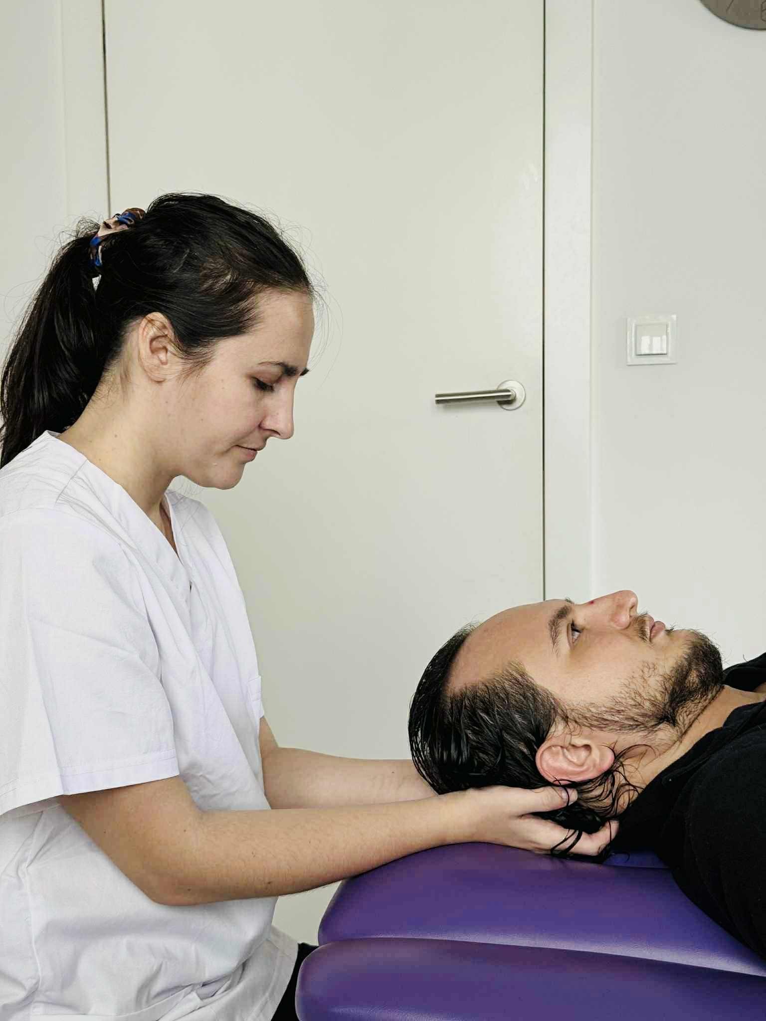 Kobieta w białym uniformie delikatnie masuje szyję pacjenta leżącego na fioletowym stole do masażu w jasnym gabinecie. Skupienie na relaksacji i terapii manualnej.