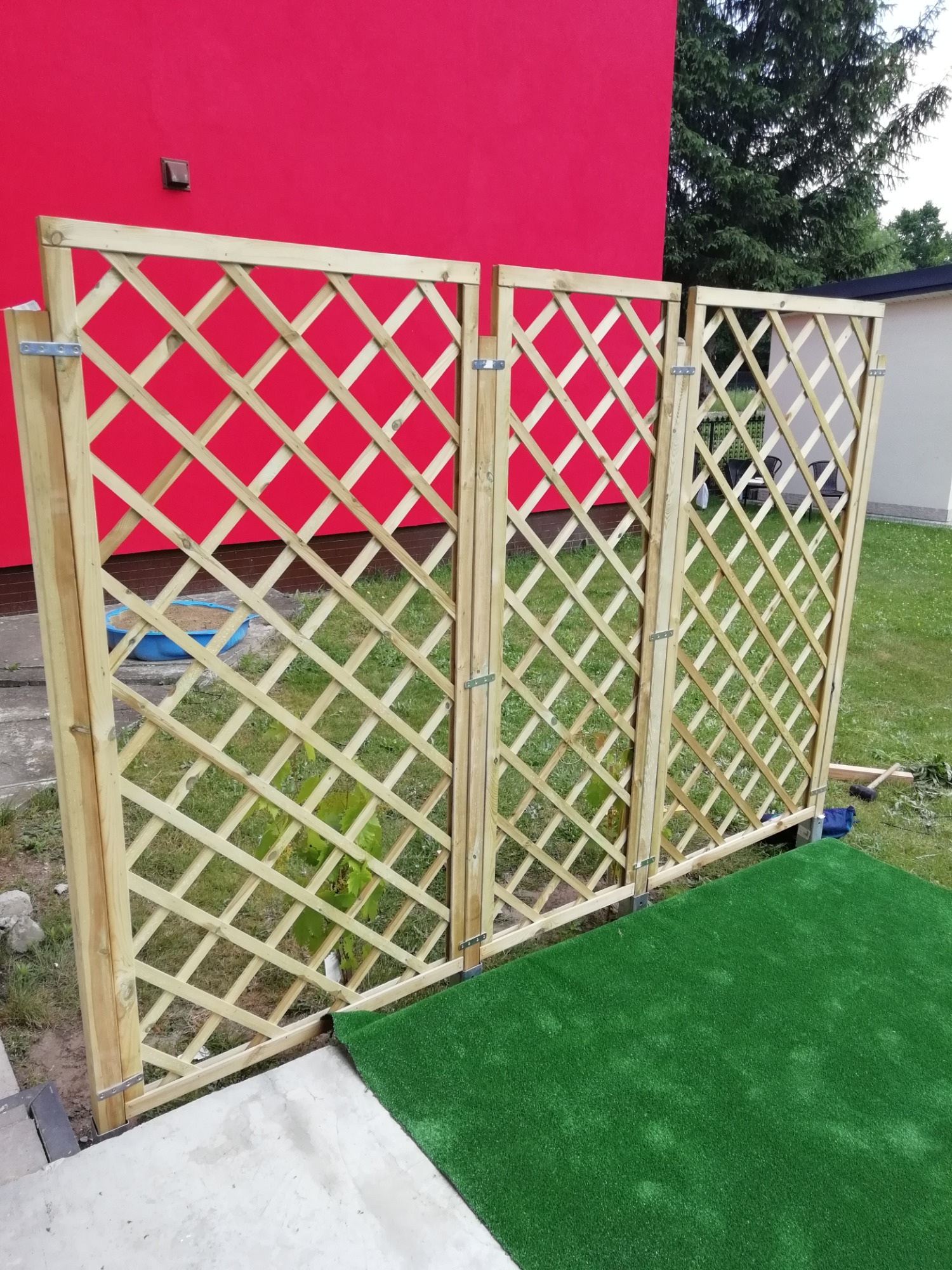 Drewniana, ażurowa osłona tarasowa z trzema segmentami, zamontowana na zewnątrz, z widoczną czerwoną ścianą budynku w tle i fragmentem sztucznej trawy na podłożu.