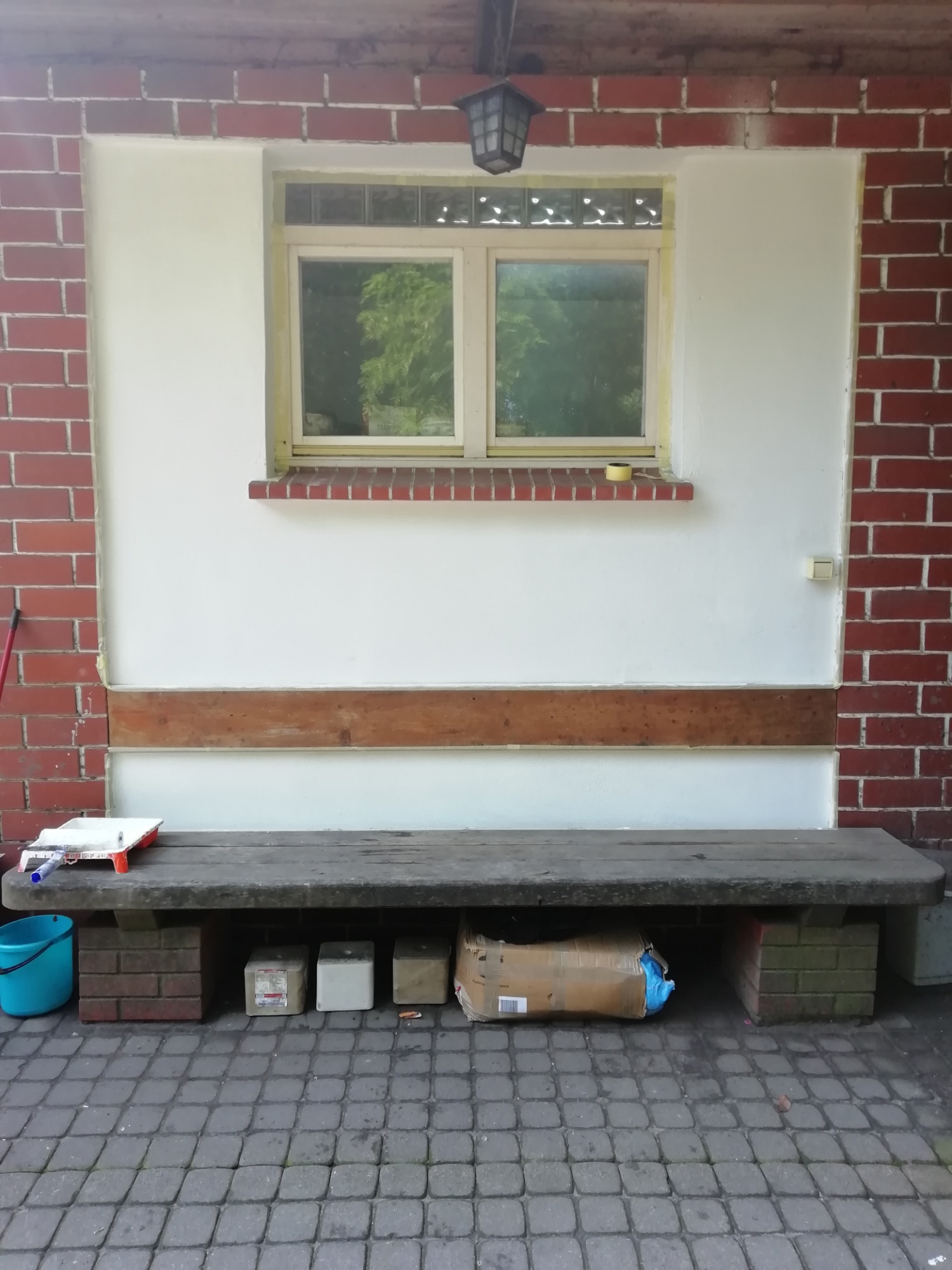 Przygotowania do odświeżenia elewacji: okno zabezpieczone taśmą, ściana częściowo pomalowana na biało, widoczny wałek i kuweta malarska na ławce.