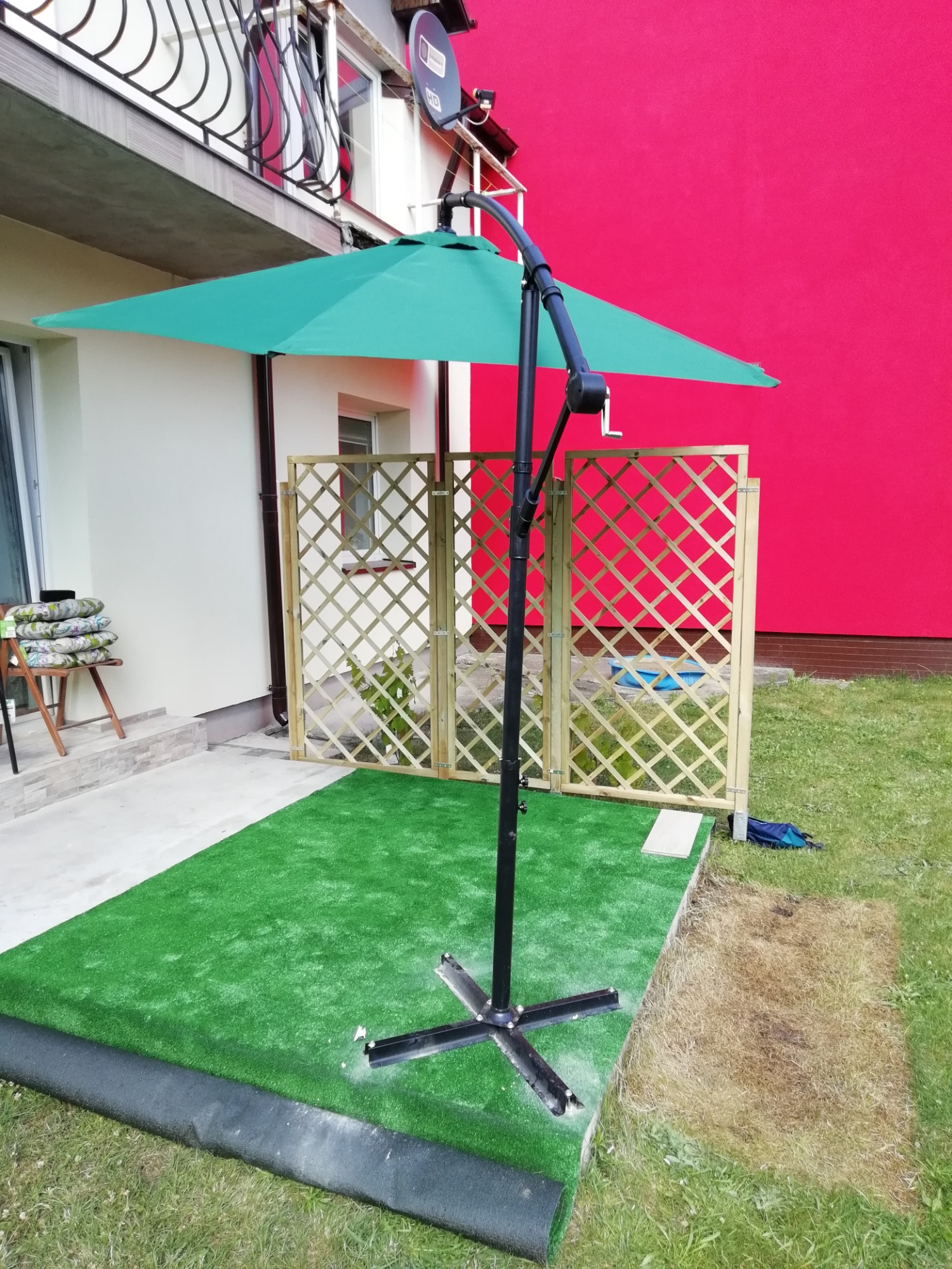 Zielony parasol ogrodowy na sztucznej trawie, drewniana ażurowa ścianka działowa, czerwona ściana budynku w tle.
