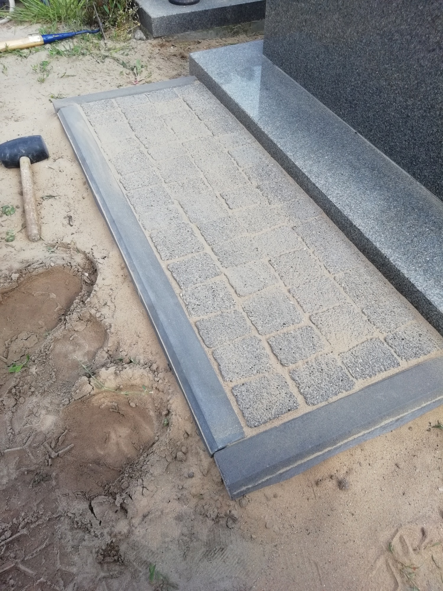 Ułożona kostka brukowa wokół nagrobka, częściowo zasypana piaskiem, obok leży młotek brukarski.