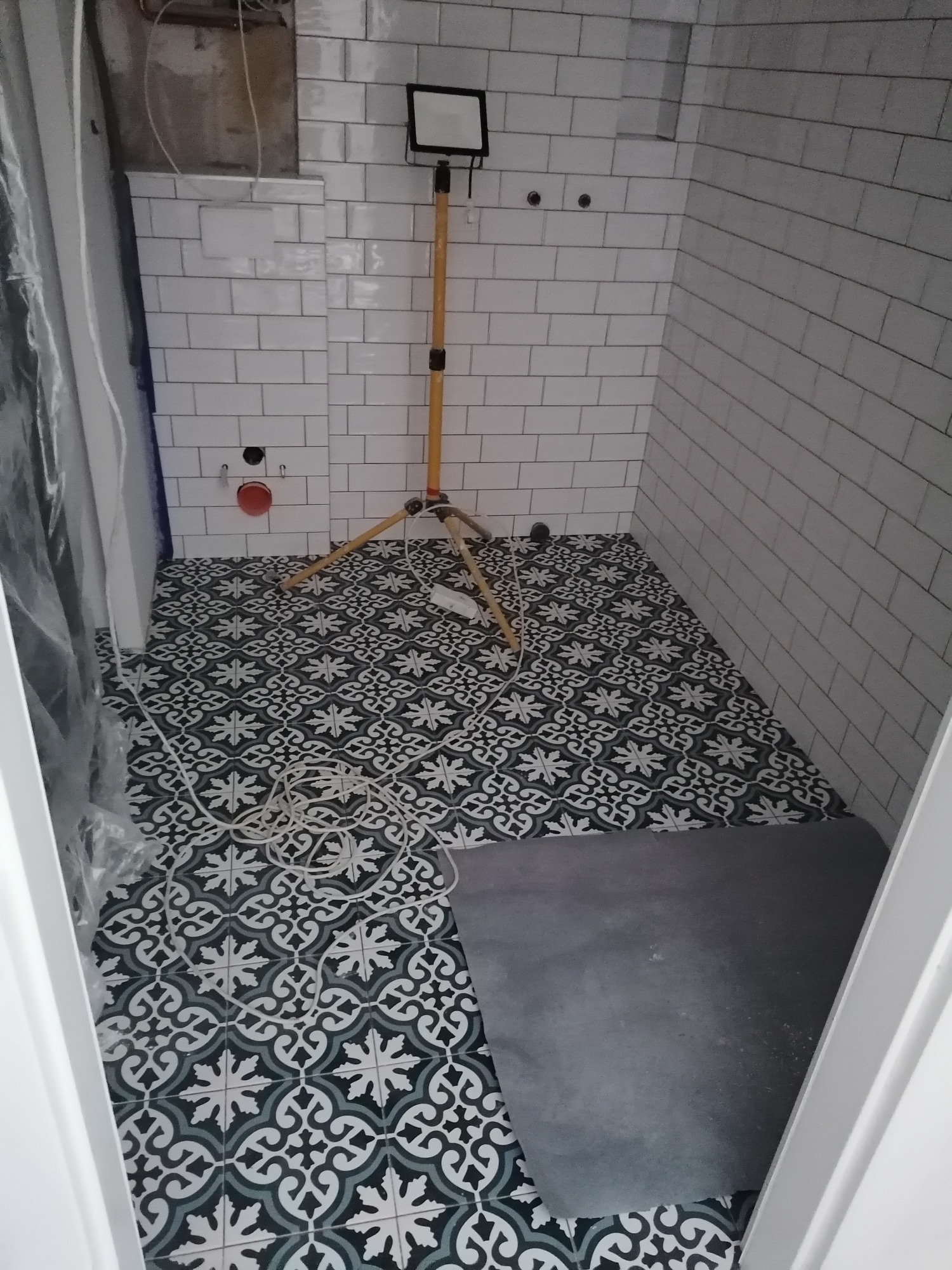 Wnętrze łazienki w trakcie prac wykończeniowych: białe płytki ścienne ułożone w cegiełkę, wzorzysta czarno-biała podłoga, fragment szarej powierzchni podłogi zabezpieczonej folią, lampa budowlana...