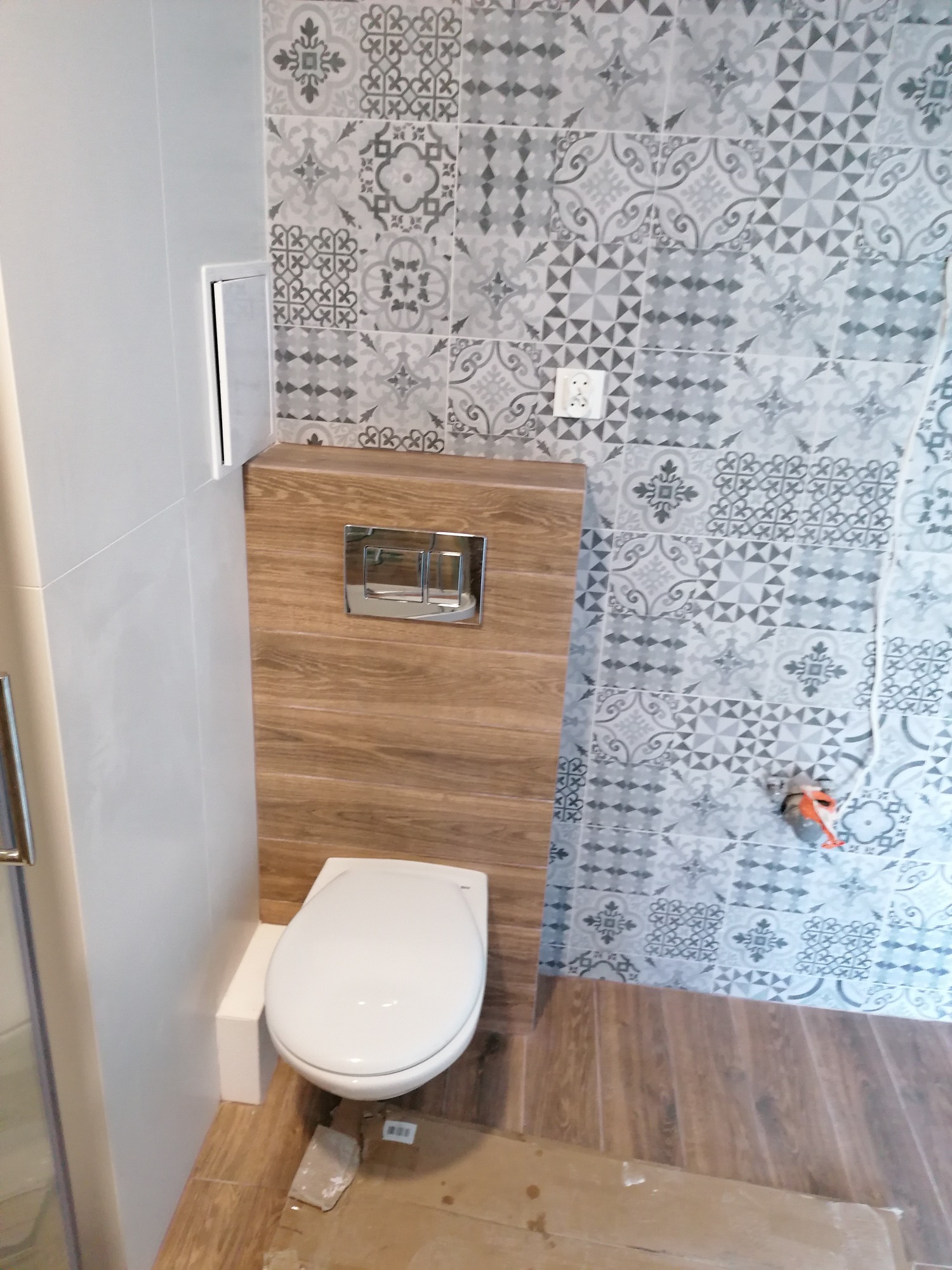 Wisząca toaleta z chromowanym przyciskiem spłukującym na tle drewnopodobnej zabudowy, ściana wyłożona dekoracyjnymi płytkami w odcieniach szarości, podłoga drewnopodobna, widoczny fragment...