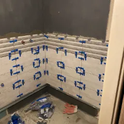 Świeżo ułożone, poziome płytki imitujące drewno na ścianie, z użyciem niebieskich krzyżyków dystansowych; widoczne puszki instalacyjne, szare ściany w górnej części i worek z krzyżykami na podłodze.
