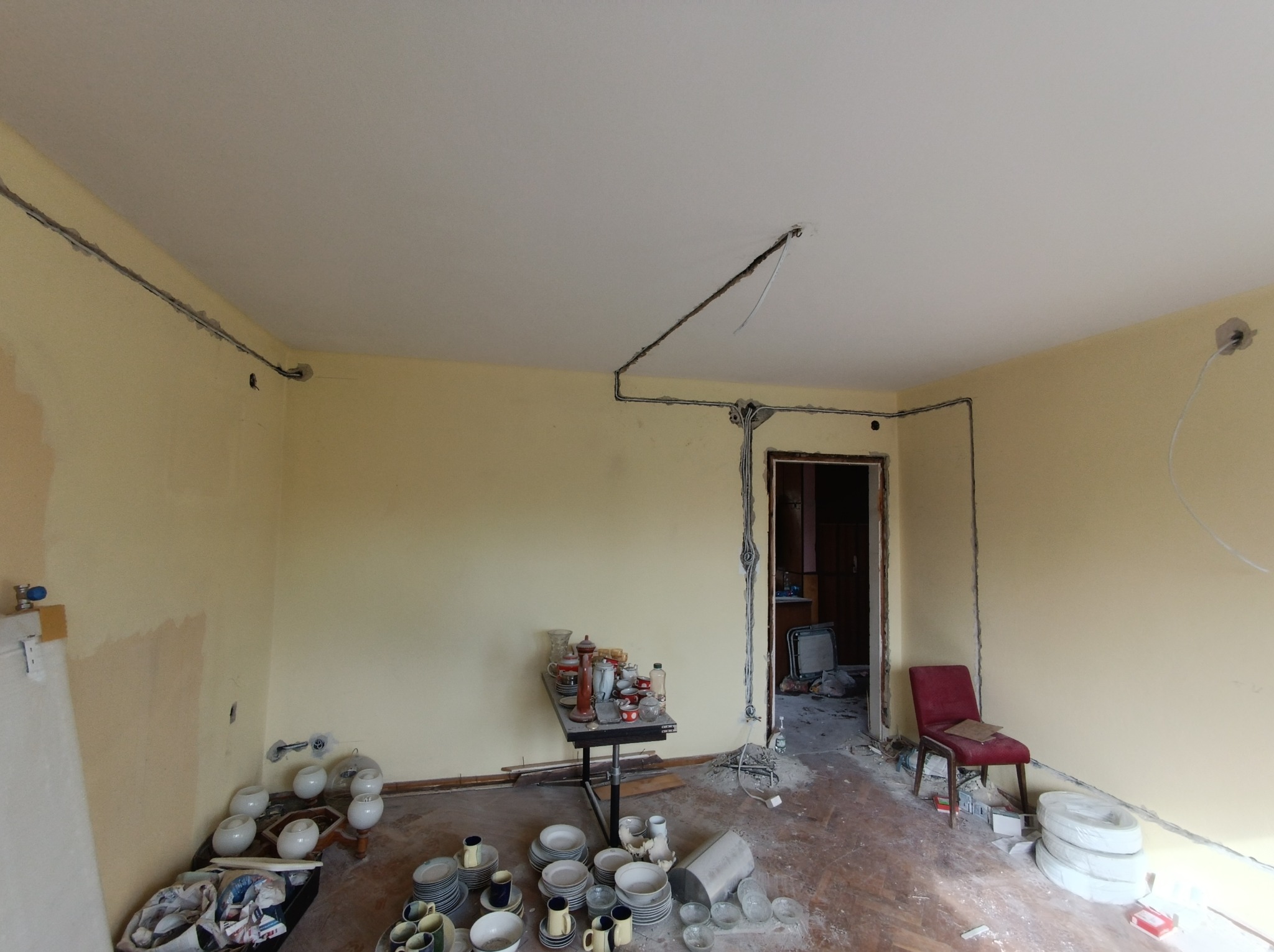 Wnętrze pokoju w trakcie remontu z poprowadzonymi przewodami elektrycznymi w bruzdach na ścianach i suficie, widoczne przygotowane kable, na podłodze porozrzucane naczynia i inne przedmioty...