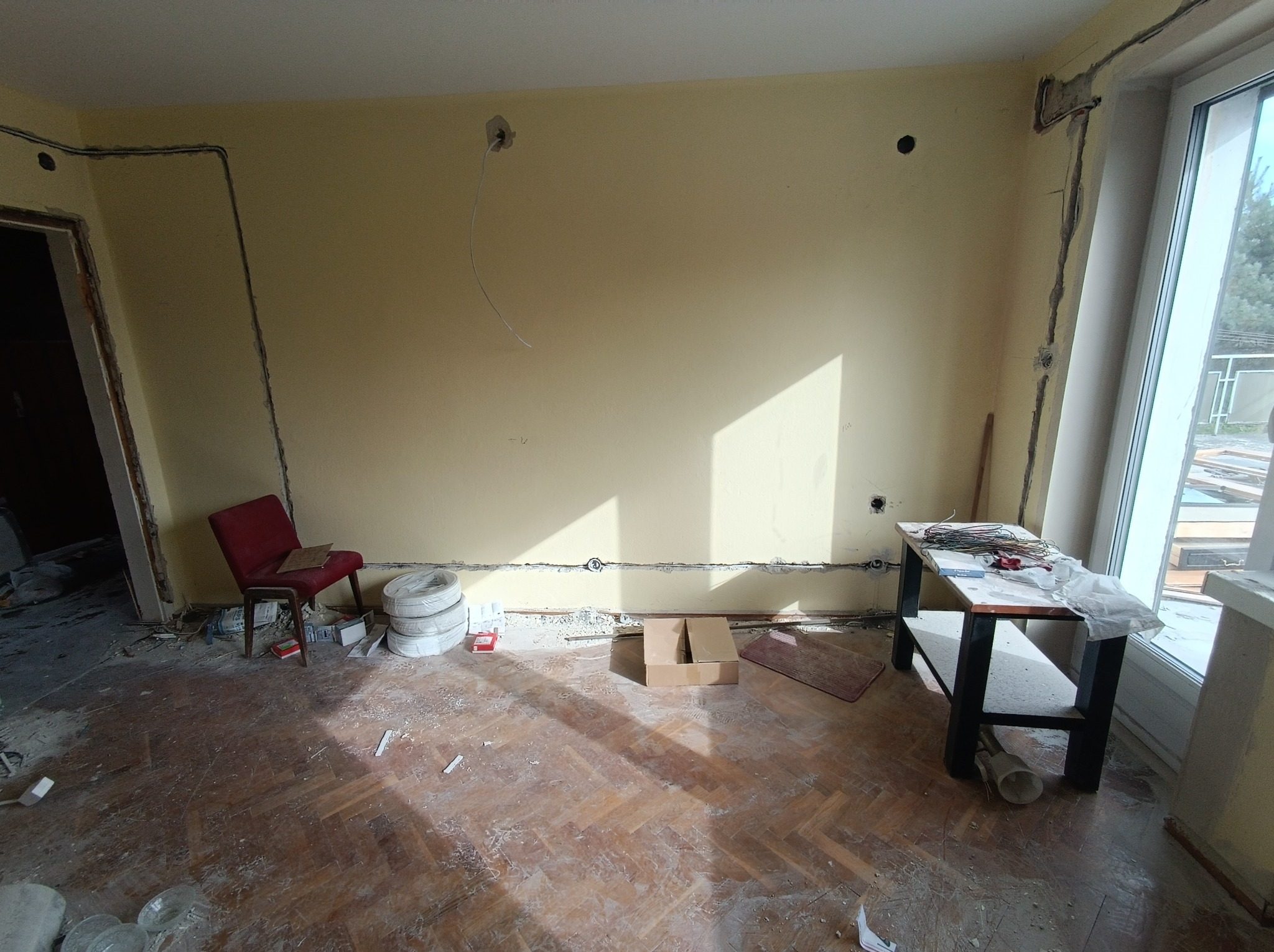 Pokój w trakcie remontu z widocznymi przewodami elektrycznymi prowadzonymi wzdłuż ścian, przygotowanymi do montażu. Na podłodze parkiet, widoczne ślady po kuciu ścian, krzesło i prowizoryczny stół...