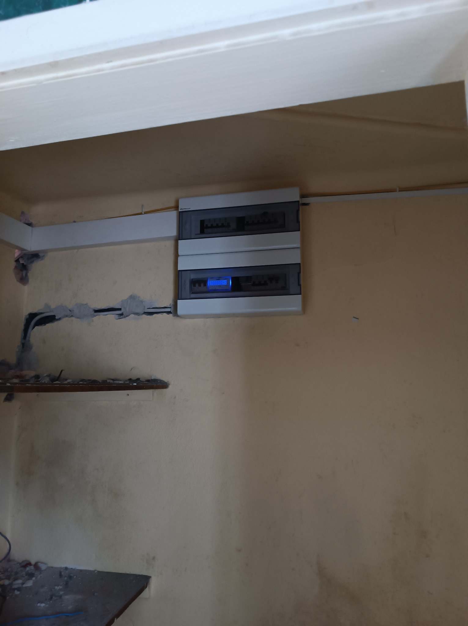 Dwu-segmentowa szara skrzynka rozdzielcza z niebieskim wyświetlaczem, zamontowana na jasnej ścianie. Widoczne przewody elektryczne w ścianie, zabezpieczone gipsem.