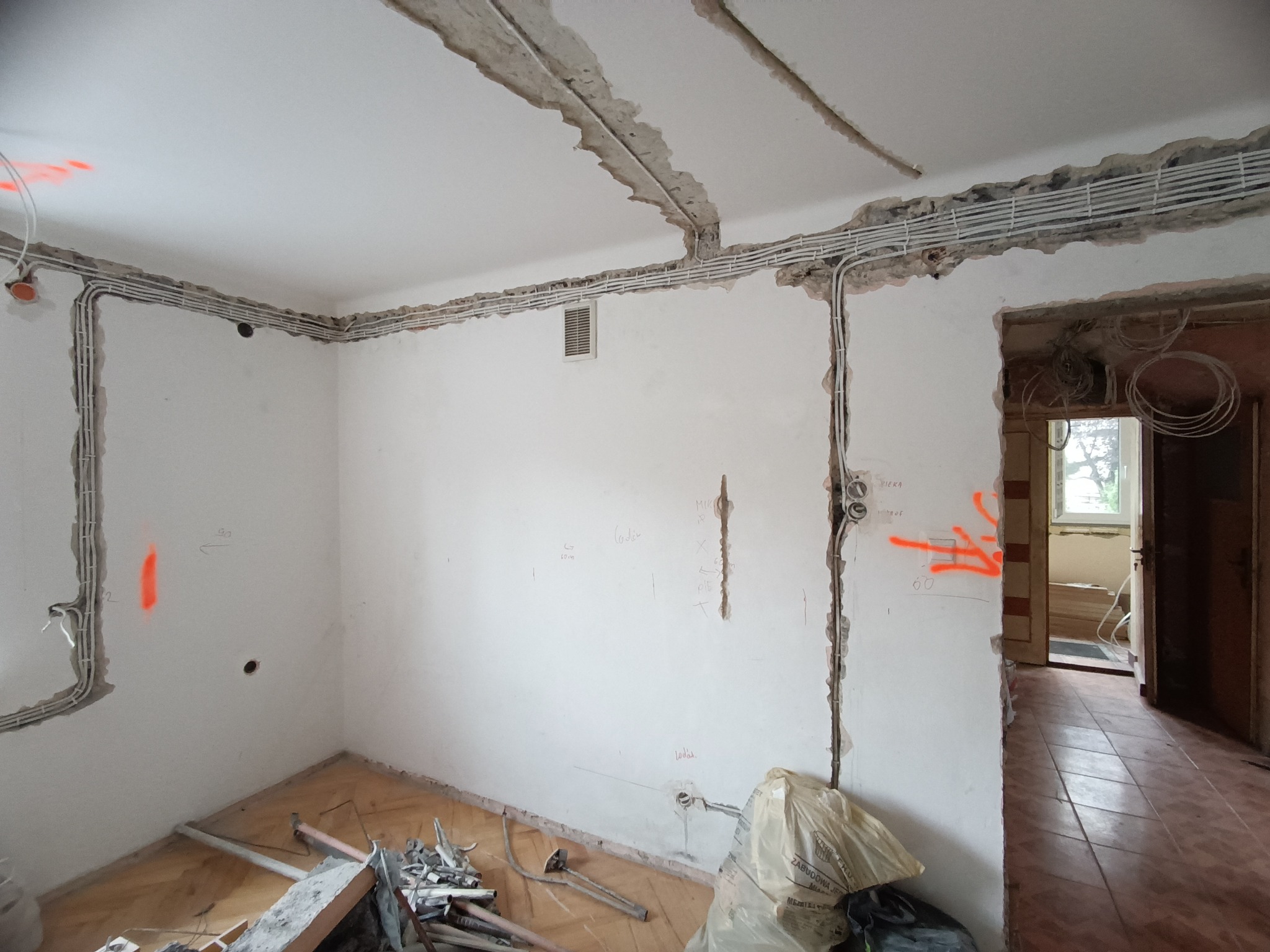 Białe kable elektryczne ułożone w bruzdach na ścianach i suficie remontowanego pokoju. Widoczne pomarańczowe oznaczenia i narzędzia na podłodze.
