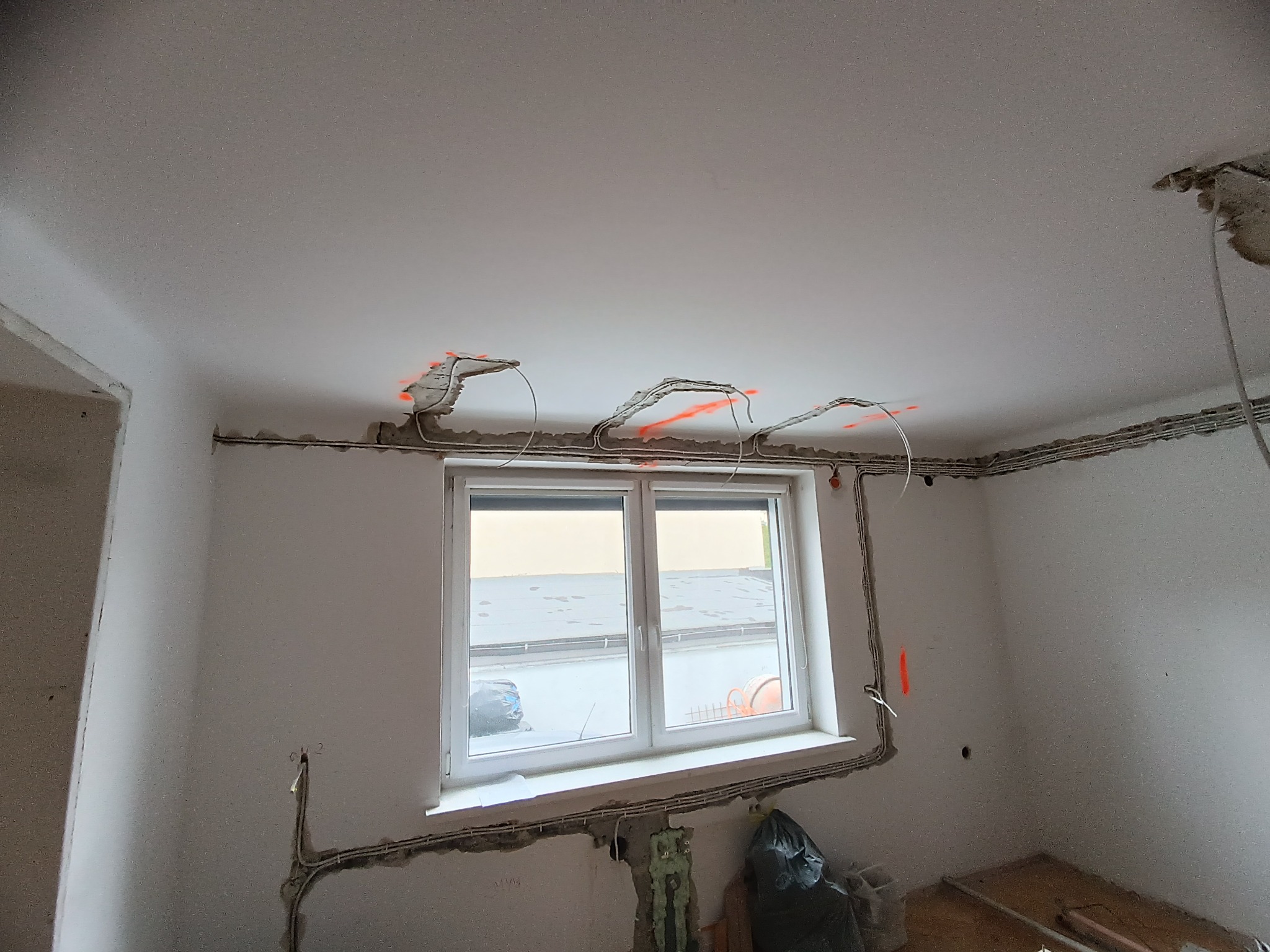 Wnętrze w trakcie remontu z widocznymi przewodami elektrycznymi ułożonymi na ścianach i suficie, oznaczonymi pomarańczowym sprayem. Widoczne okno i surowy stan wykończenia.
