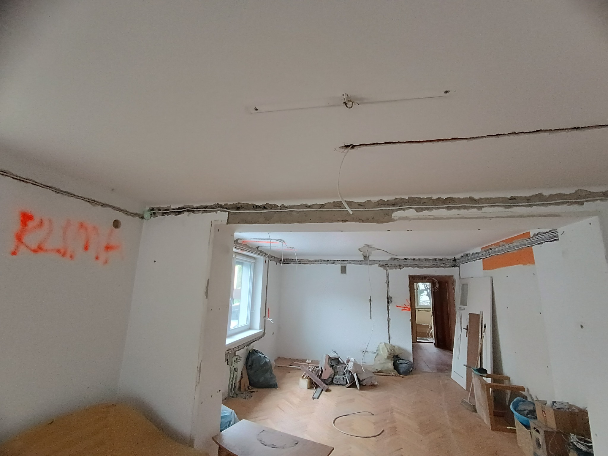 Wnętrze w trakcie remontu z widocznymi bruzdami pod instalacje elektryczne na ścianach i suficie. Na ścianie pomarańczowy napis 'KLIMA'.