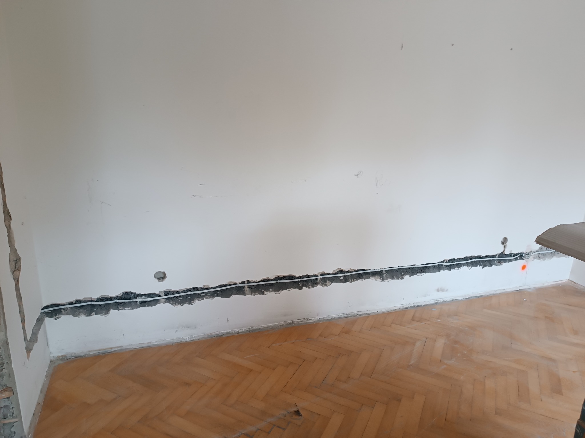 Biała ściana z wyżłobionym korytem i poprowadzonymi przewodami elektrycznymi. Parkiet w jodełkę na podłodze. Widoczne ślady po kuciu.