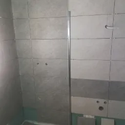 Ściana w trakcie układania płytek ceramicznych w odcieniach szarości, widoczne otwory na instalacje i listwa wykończeniowa, dolne rzędy częściowo nieukończone.