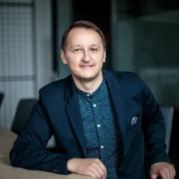 Uśmiechnięty mężczyzna w granatowej marynarce i wzorzystej koszuli siedzi przy biurku, w tle widoczne biurowe przeszklenia i krzesła.