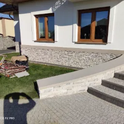 Wykończona elewacja domu z kamienną okładziną, widoczne okna z brązowymi ramami, schody wejściowe z kostki brukowej oraz przygotowane rusztowanie na trawniku.