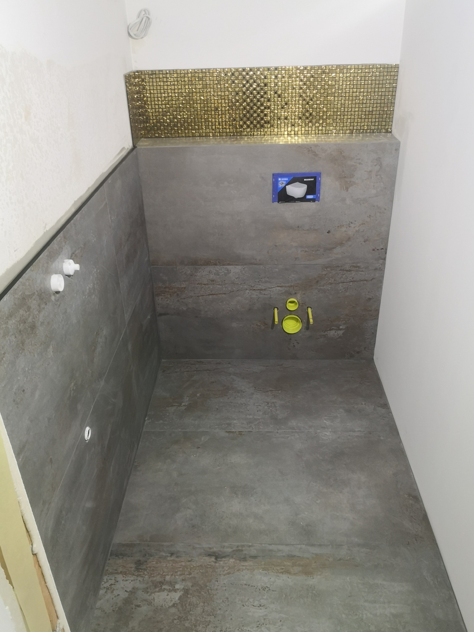 Wnętrze łazienki w trakcie remontu: szare płytki imitujące beton na ścianach i podłodze, złota mozaika powyżej stelaża podtynkowego WC, widoczne przyłącza wodno-kanalizacyjne.