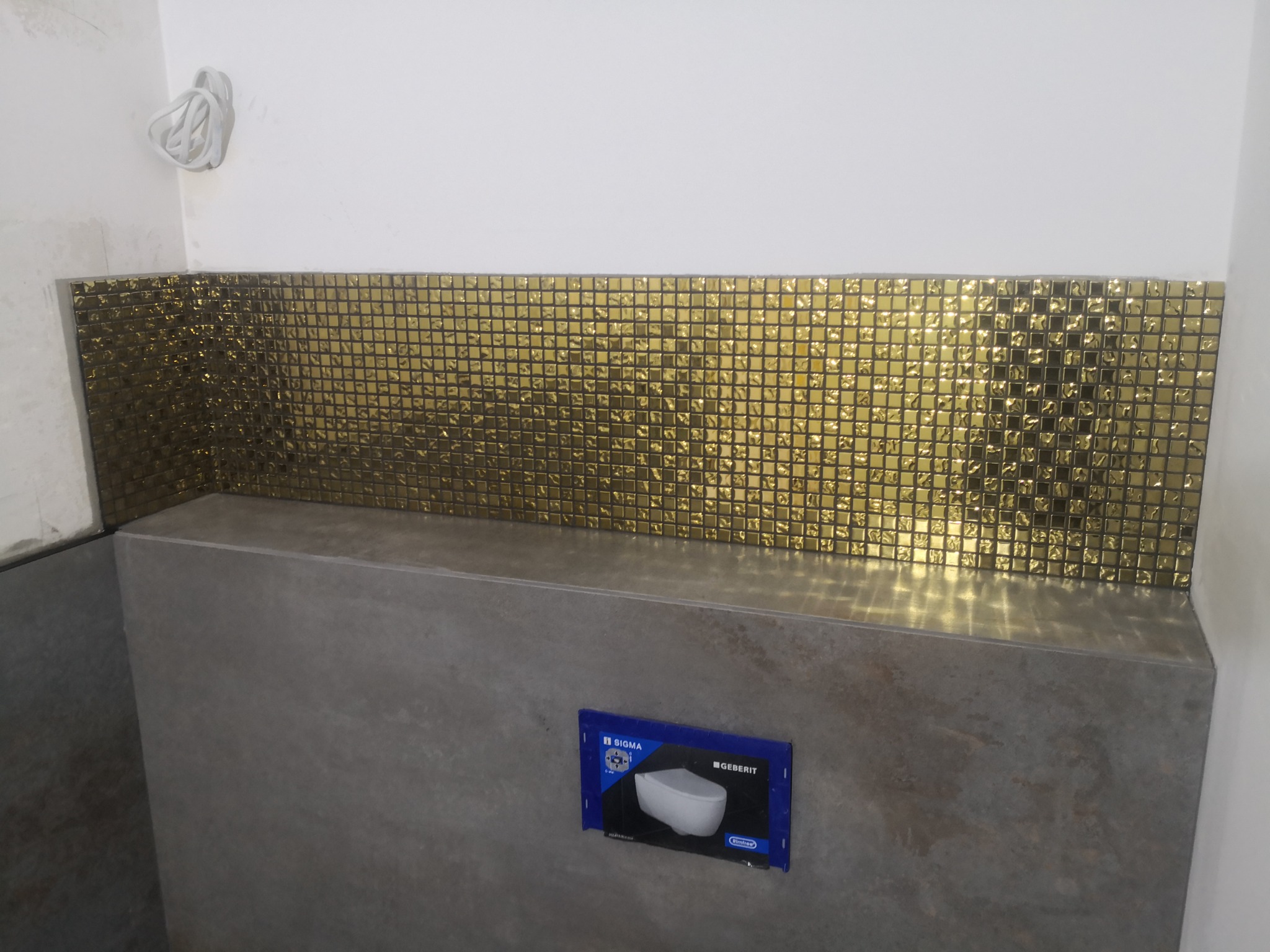 Fragment łazienki w trakcie wykańczania: złota mozaika ścienna nad zabudową stelaża podtynkowego, widoczna instalacja elektryczna.