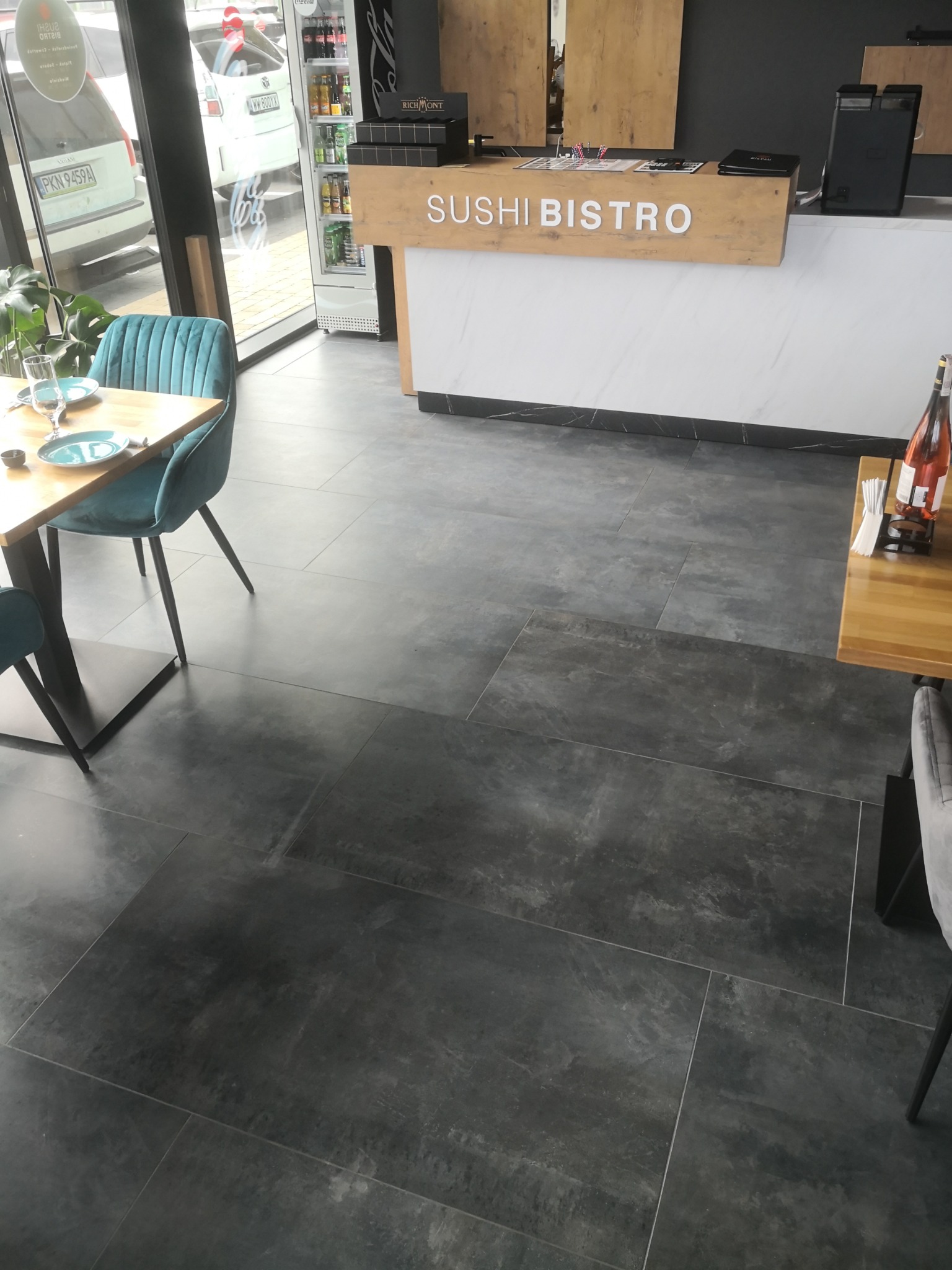 Ciemnoszara, matowa podłoga z dużych, prostokątnych płytek w lokalu Sushi Bistro, z widocznymi stolikami i krzesłami w tle.