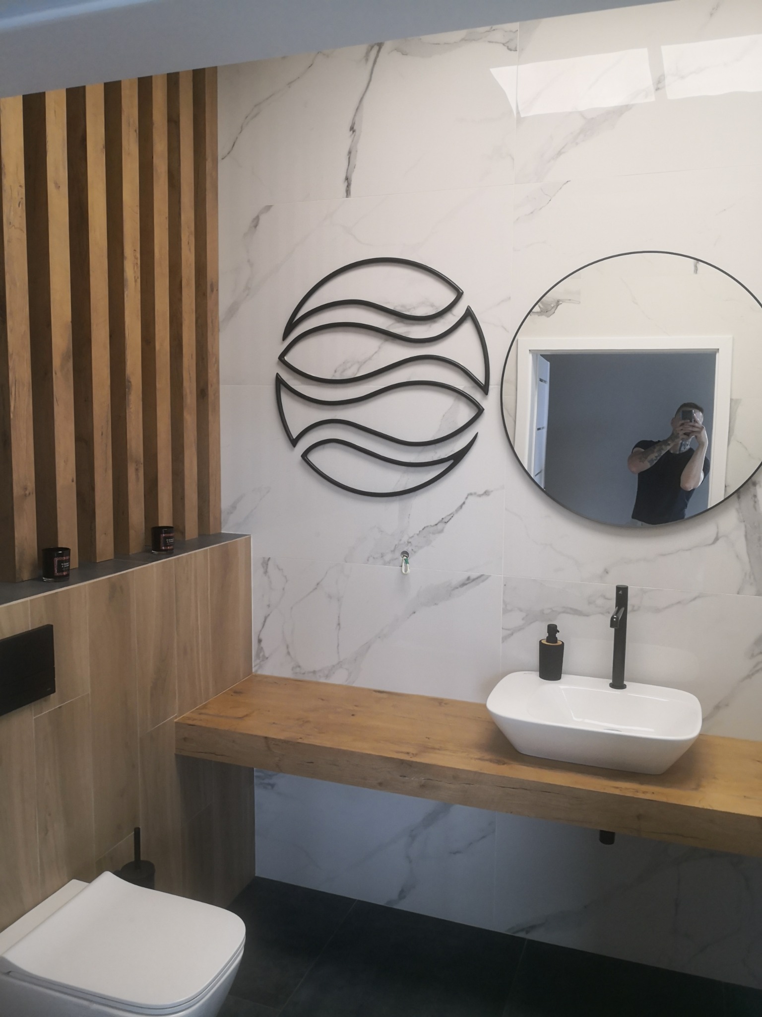 Nowoczesna łazienka z marmurowymi płytkami, drewnianą zabudową umywalki i dekoracyjnym elementem ściennym w kształcie fal, odbicie fotografa w okrągłym lustrze.