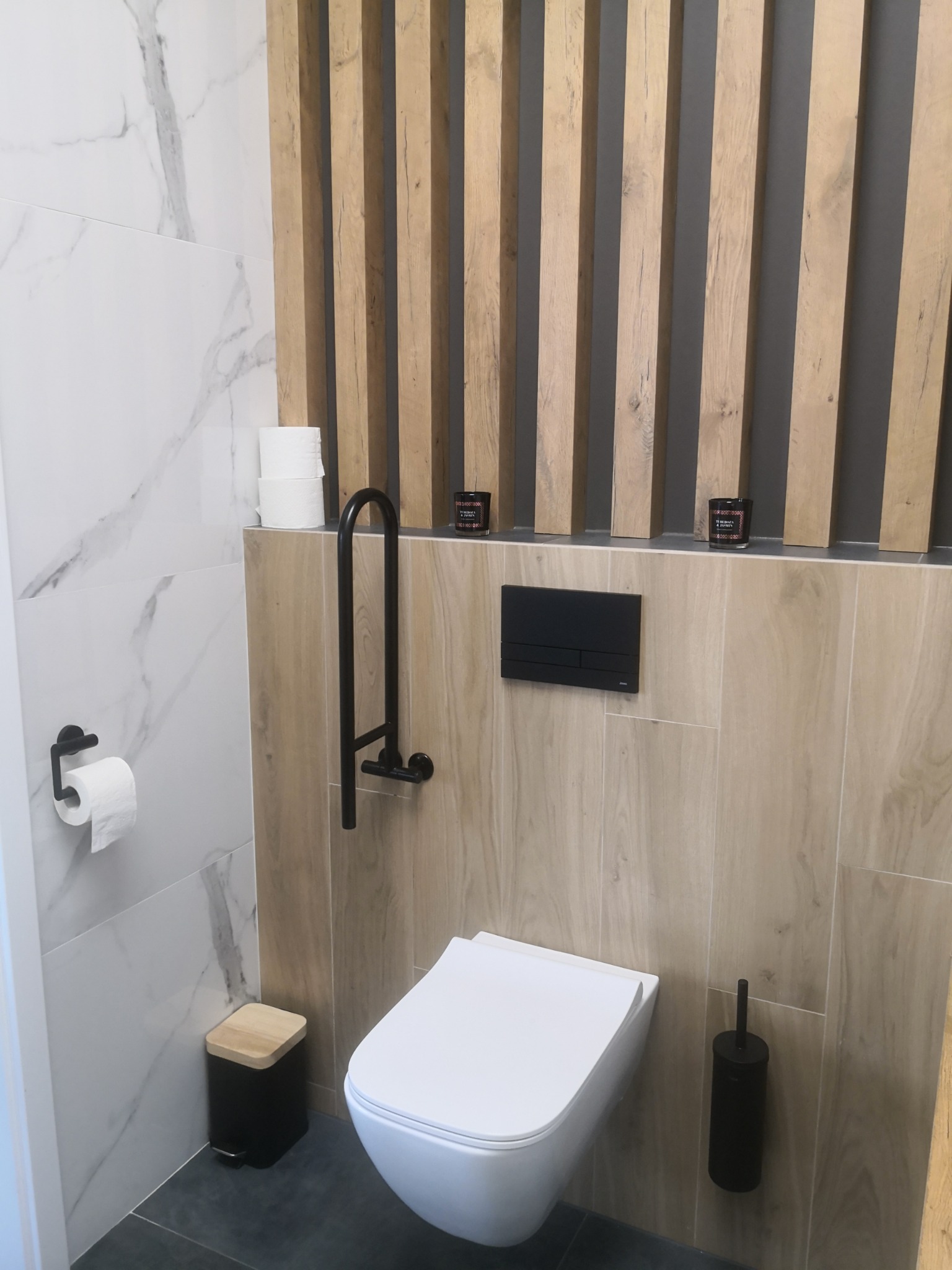 Nowoczesna łazienka z wiszącą toaletą, drewnianymi lamelami na ścianie i czarnymi akcesoriami, takimi jak uchwyt dla niepełnosprawnych, kosz na śmieci i szczotka do WC.