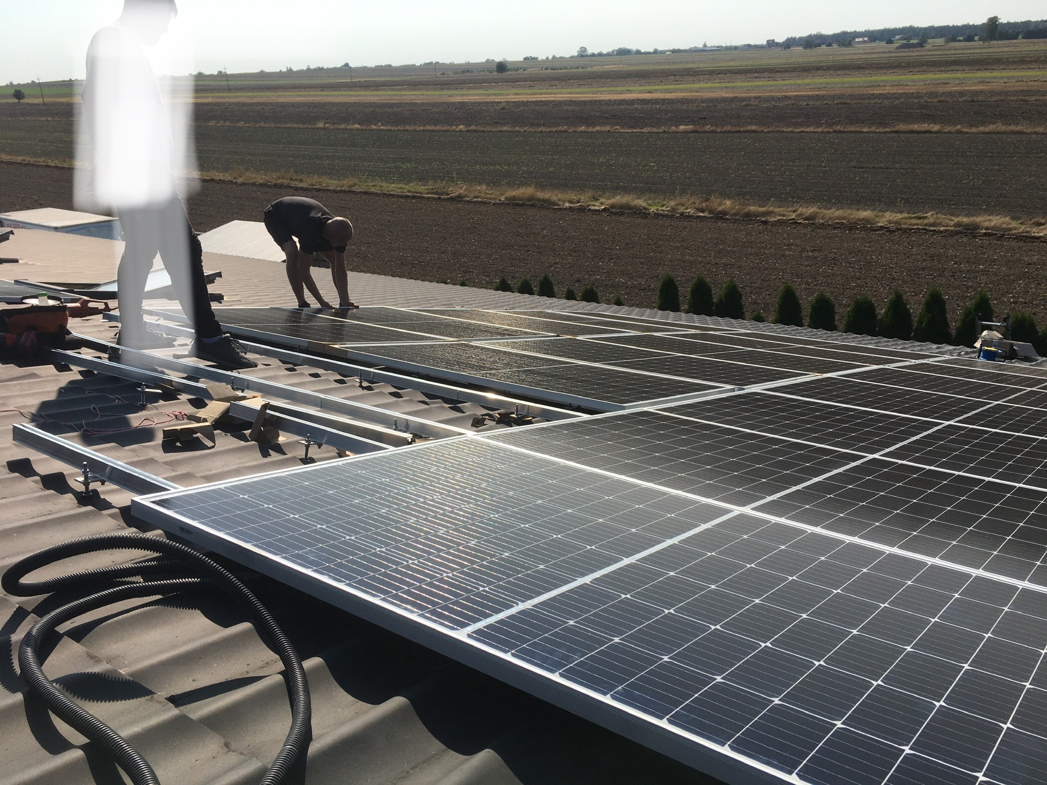 Montaż paneli fotowoltaicznych na dachu z widocznym krajobrazem pól uprawnych w tle.